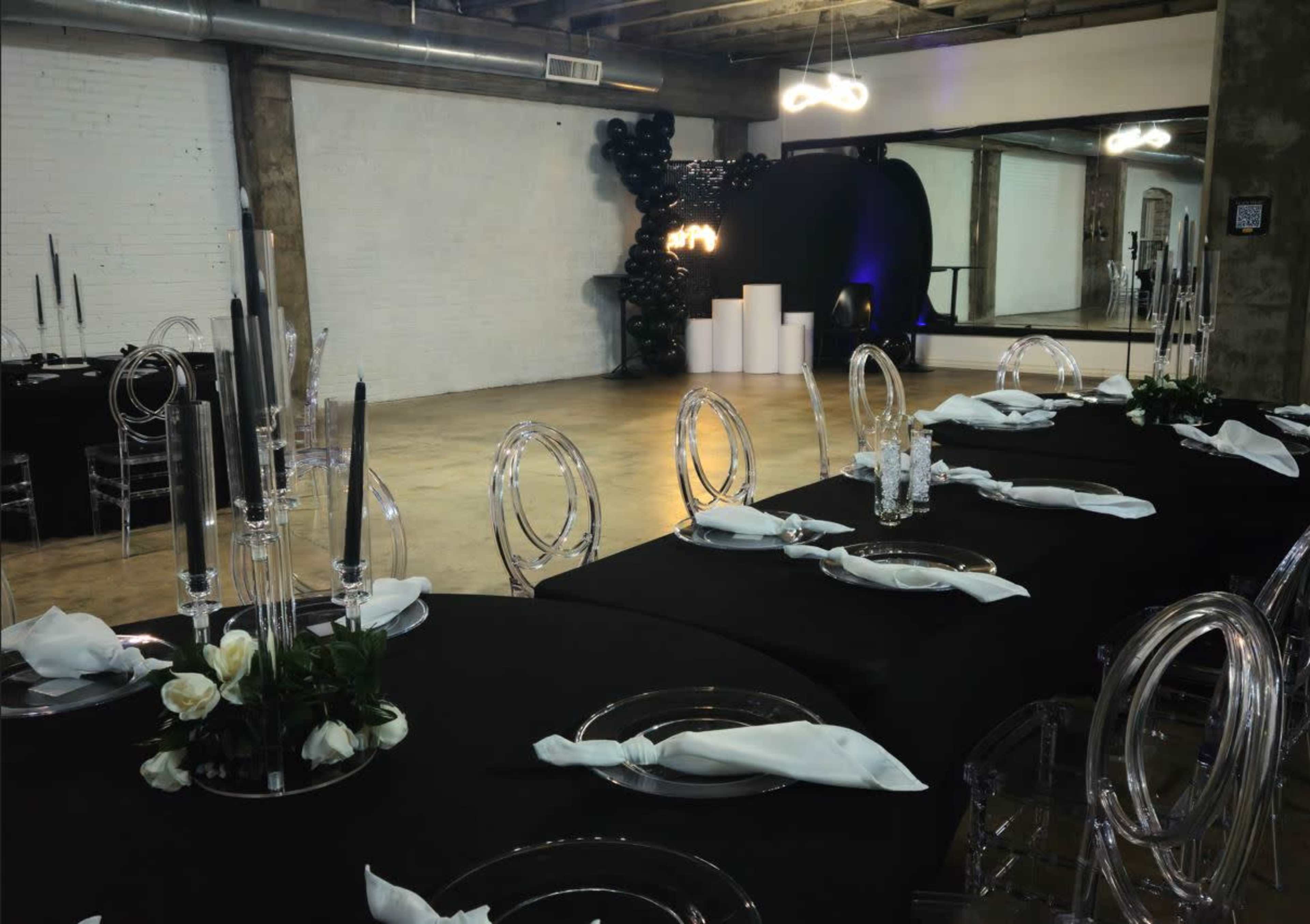 A long table is set with clear chairs and black tablecloths, adorned with white napkins and candles, in a spacious venue with a modern backdrop.