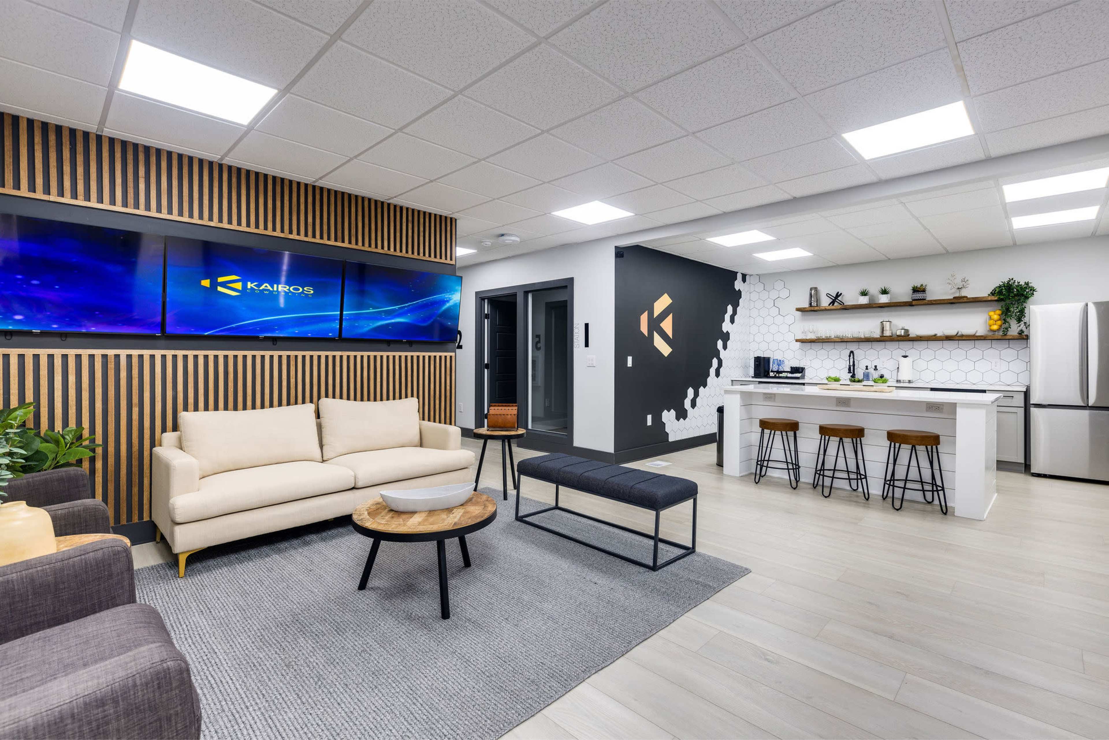 A modern office lounge area with a beige sofa, a black bench, a wooden coffee table, and a kitchen space featuring bar stools and a large screen monitor on the wall.
