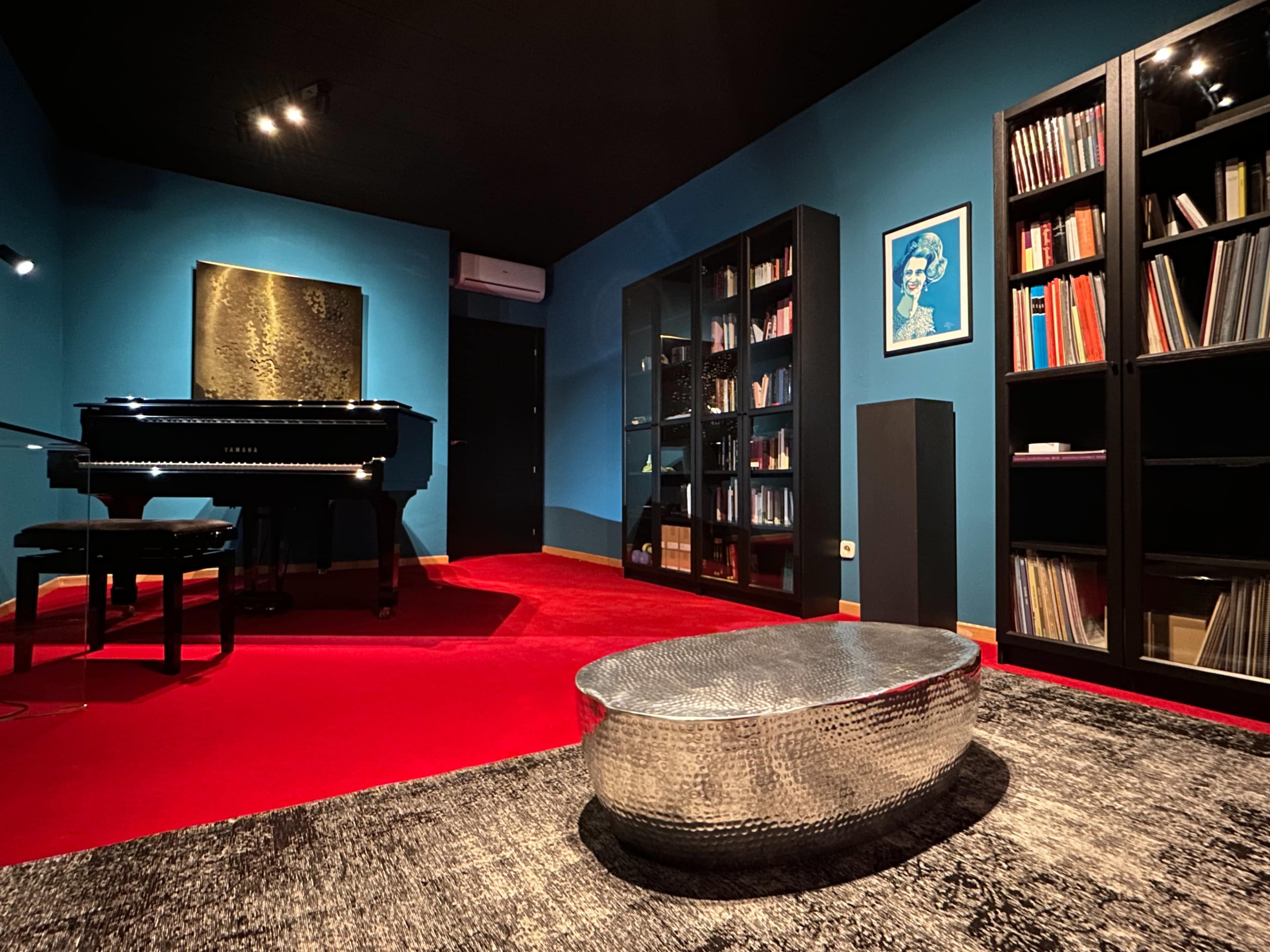A room features a grand piano, black bookshelves filled with books, and a metallic coffee table on a red carpet against teal walls.