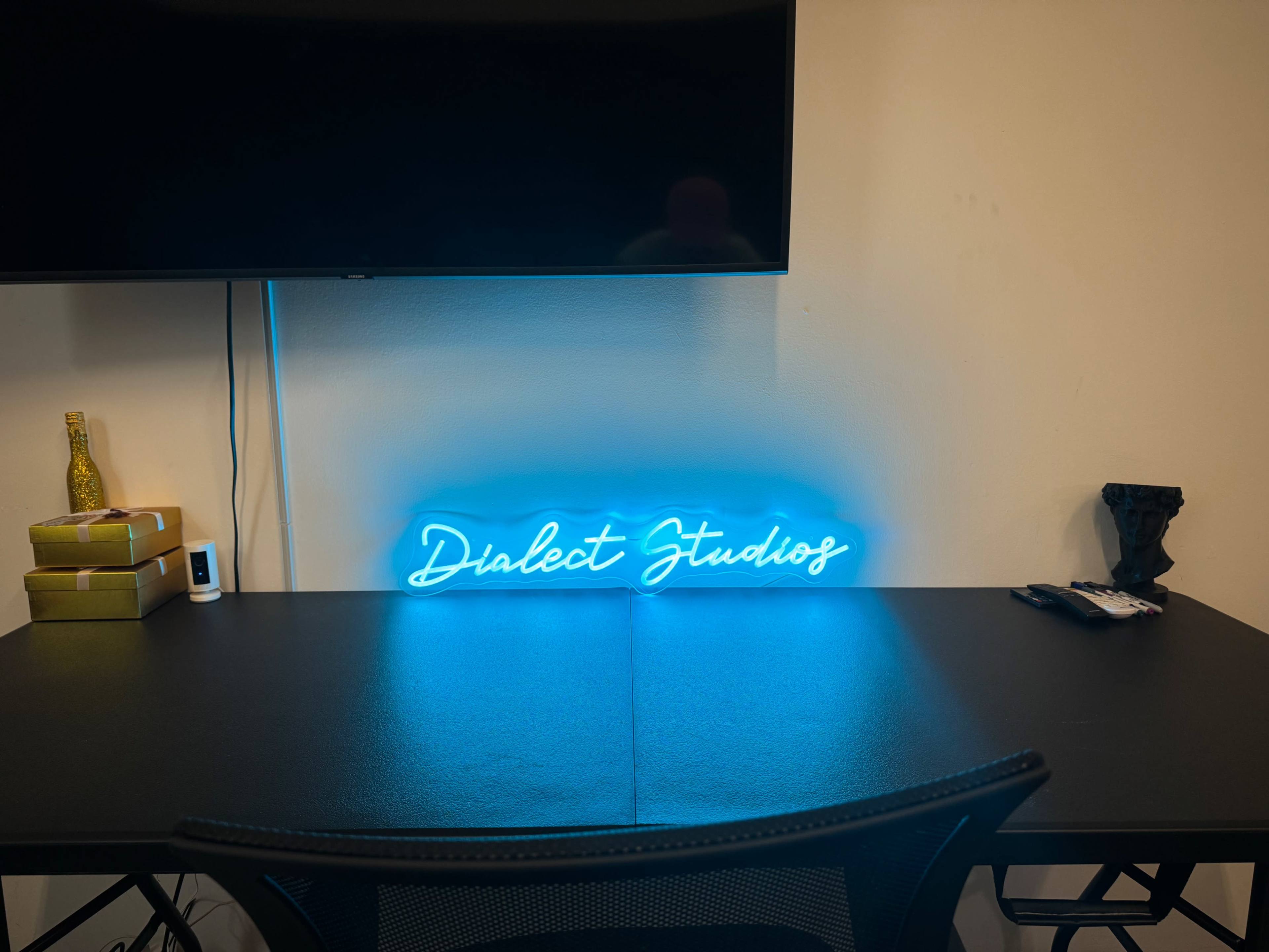 The image shows a desk with a neon sign that reads "Dialect Studios," a television mounted above it, and various items including books and a decorative object on the surface.