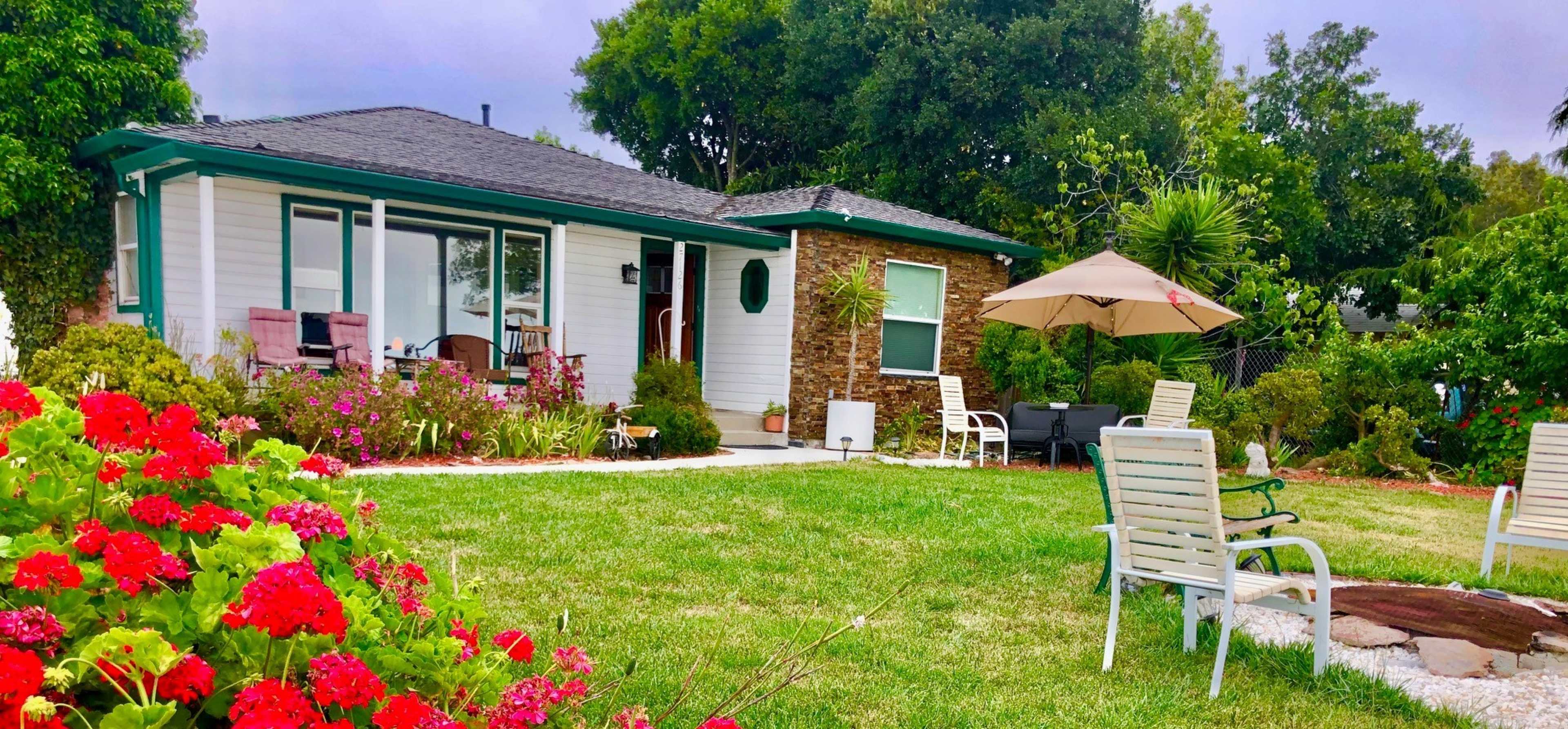 A single-story house with a garden featuring colorful flowers, a patio umbrella, and outdoor seating arrangements.