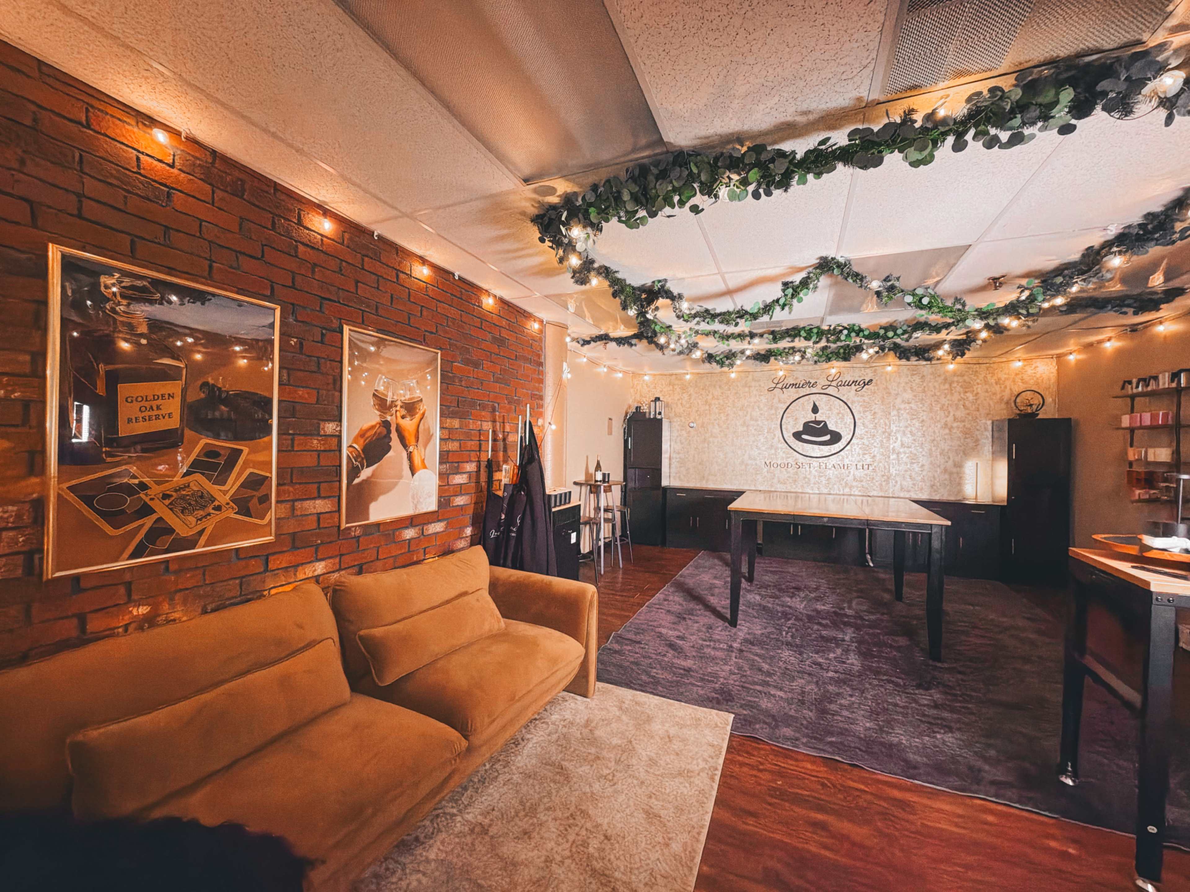 The image shows a cozy interior space with a brick wall, festive string lights, and a large table set for activities, accompanied by framed art on the walls.