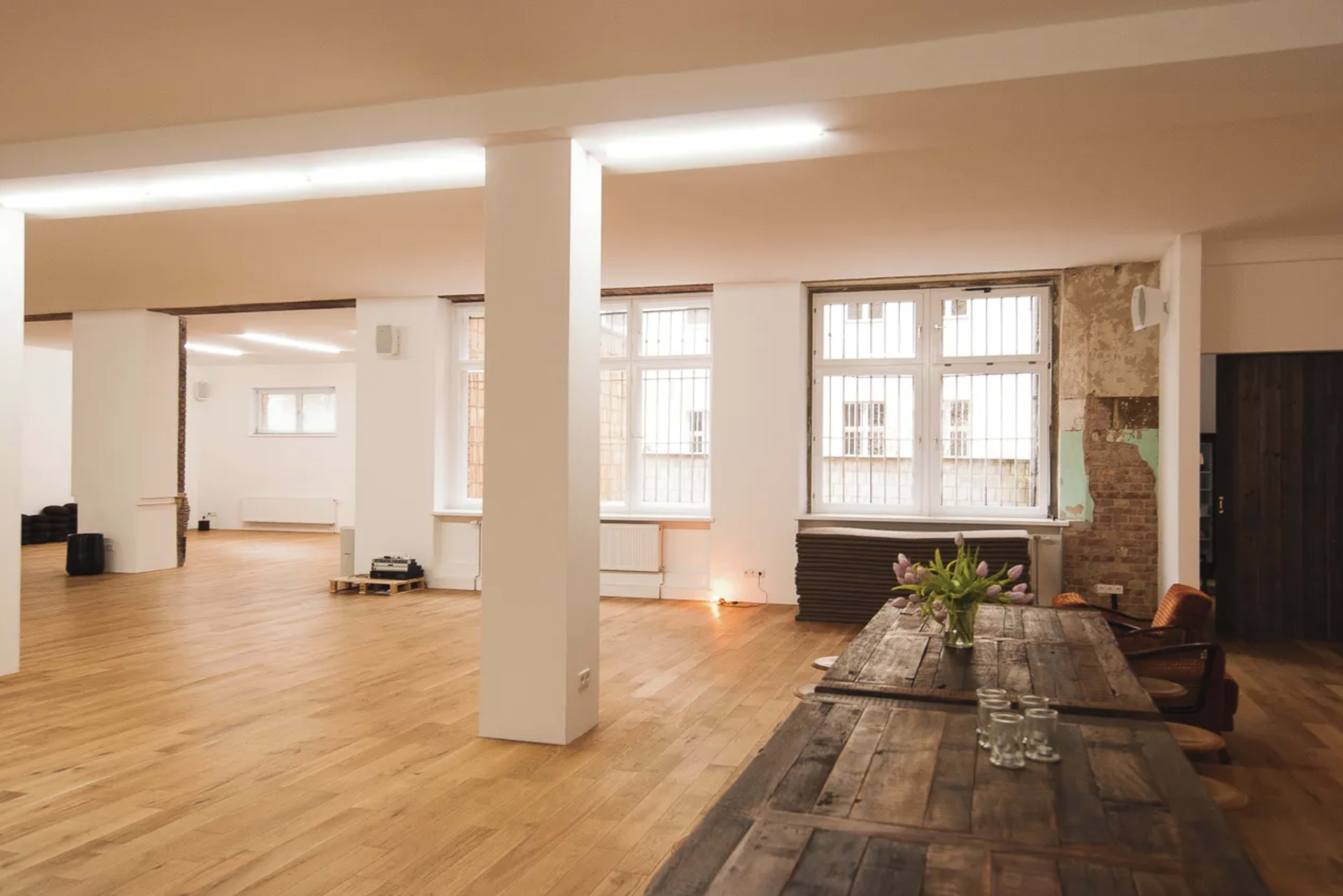 A spacious, minimalist room with wooden floors, large windows, and a long wooden table set with glasses.