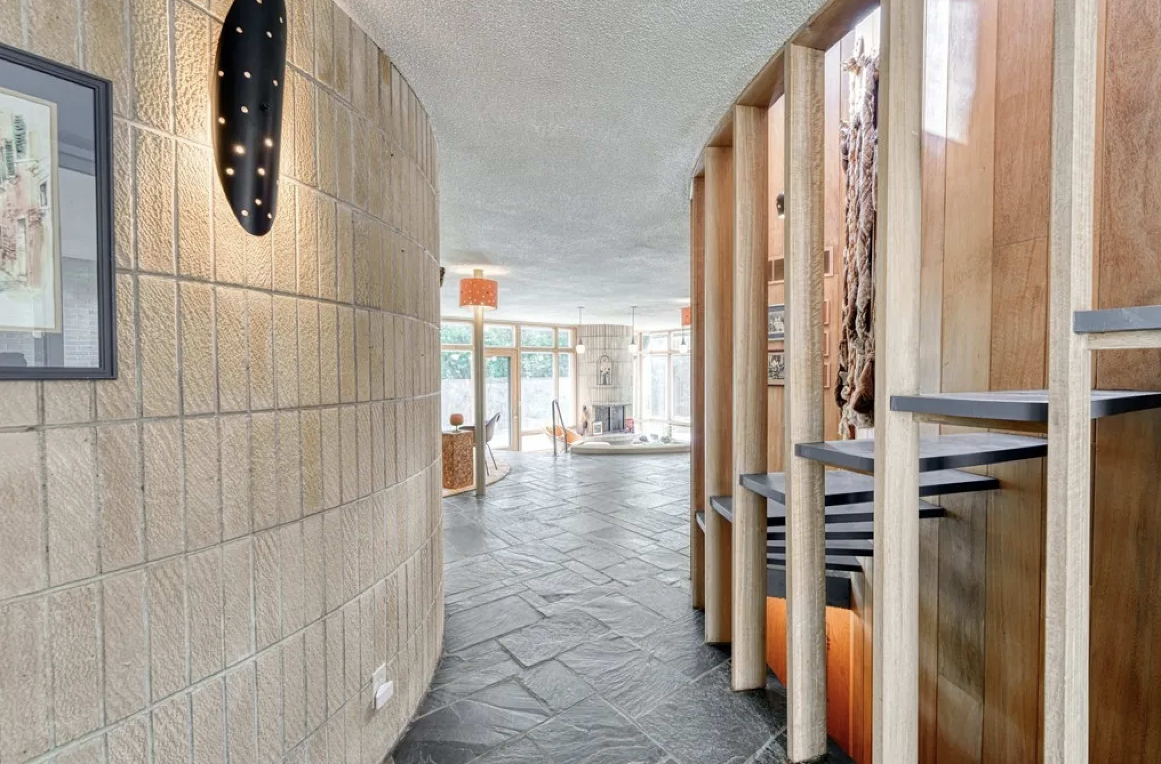A curved hallway with textured walls and wooden accents leads to a brightly lit living area with large windows and modern furnishings.