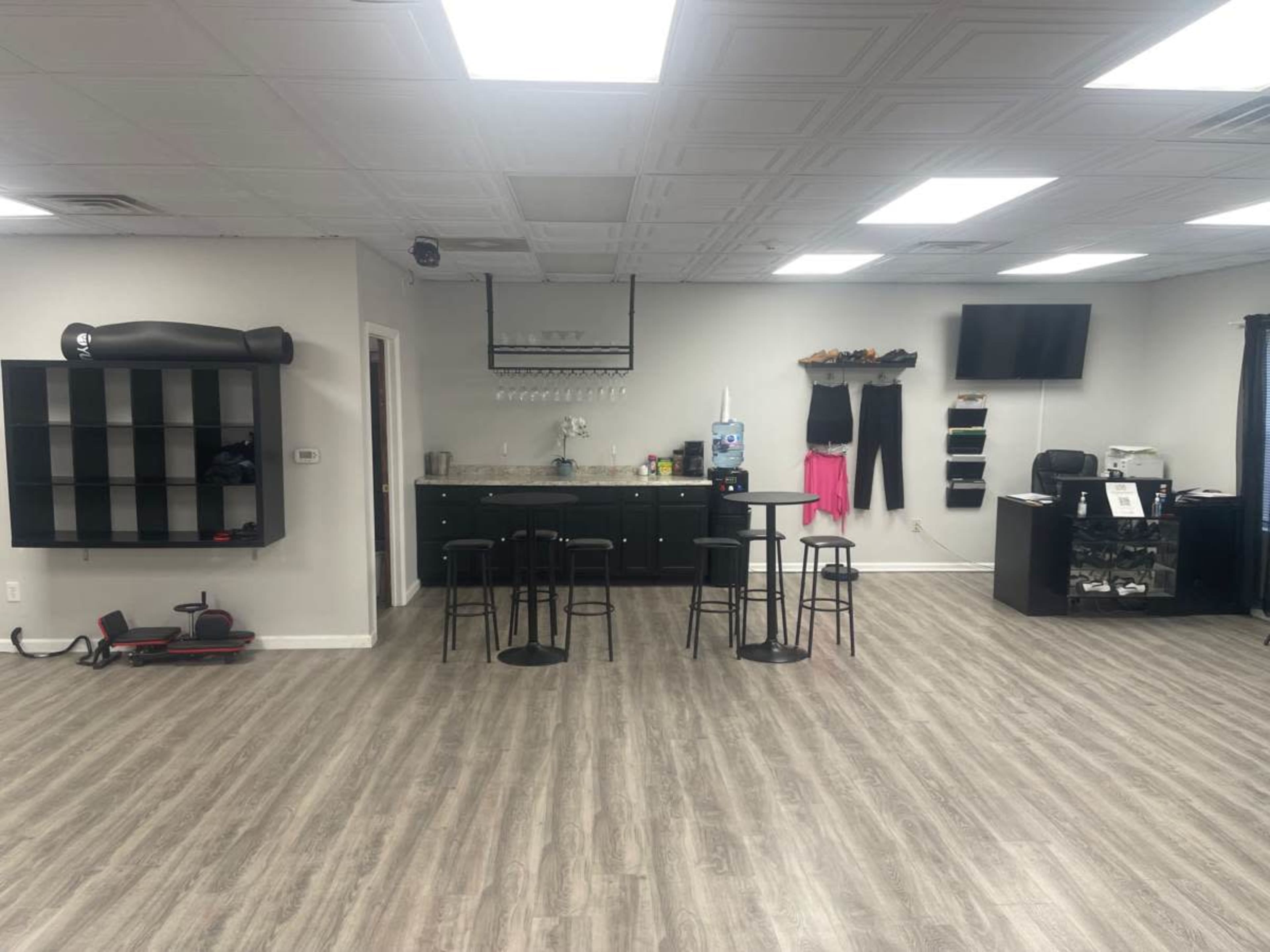 A spacious, well-lit room with a small kitchenette area, black barstools, a desk with office supplies, and workout equipment along the walls.