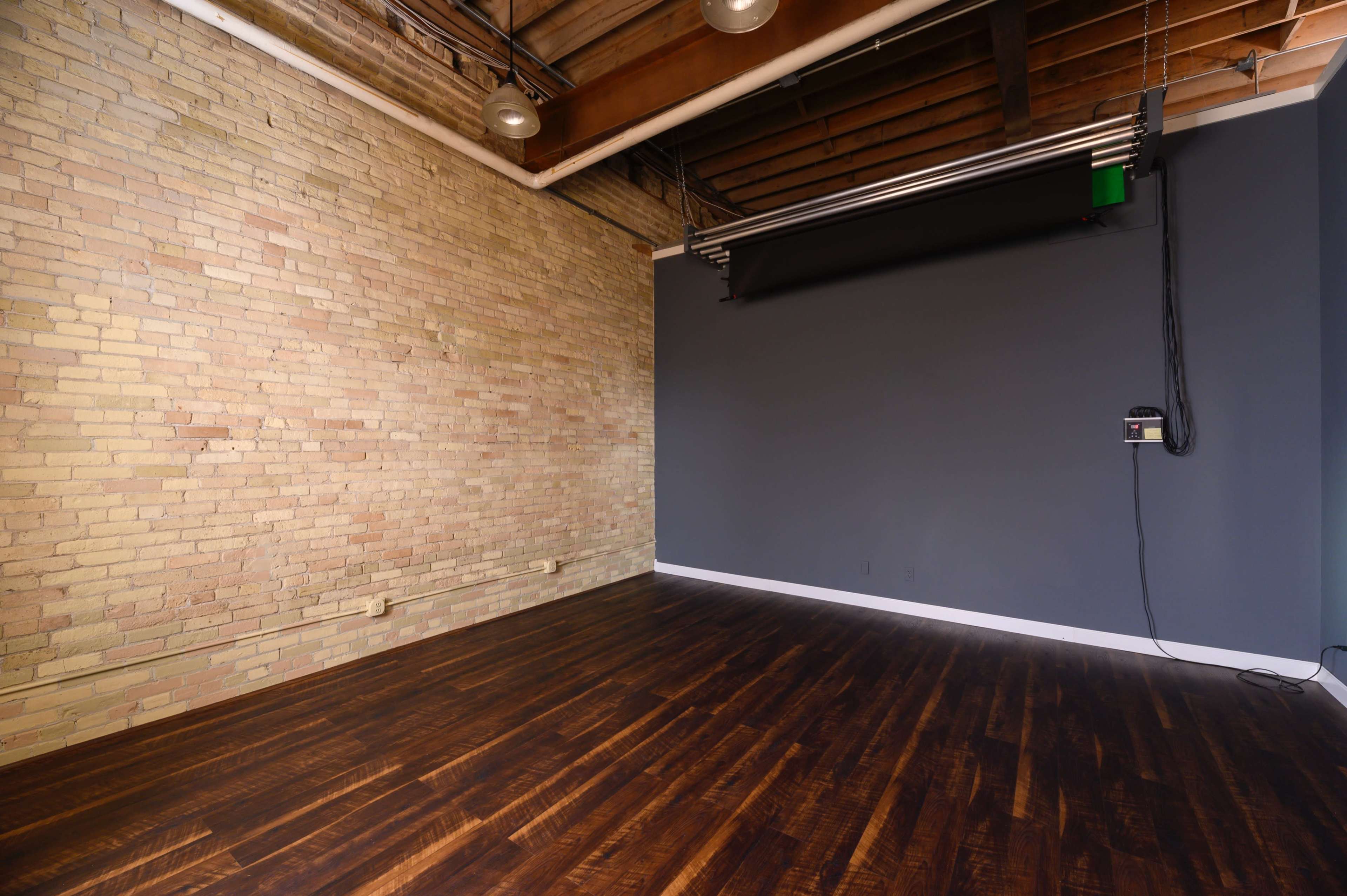 A spacious room with exposed brick walls, dark wooden flooring, and a dark gray wall with a roll-down screen.