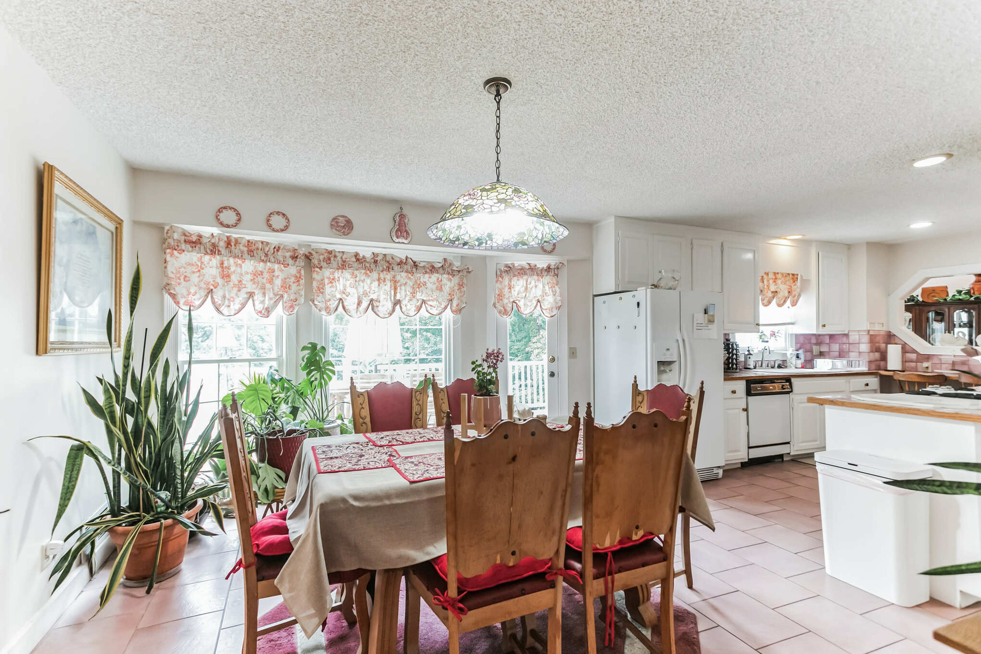 A bright kitchen with a dining table surrounded by wooden chairs, large windows adorned with floral curtains, and various plants in pots.