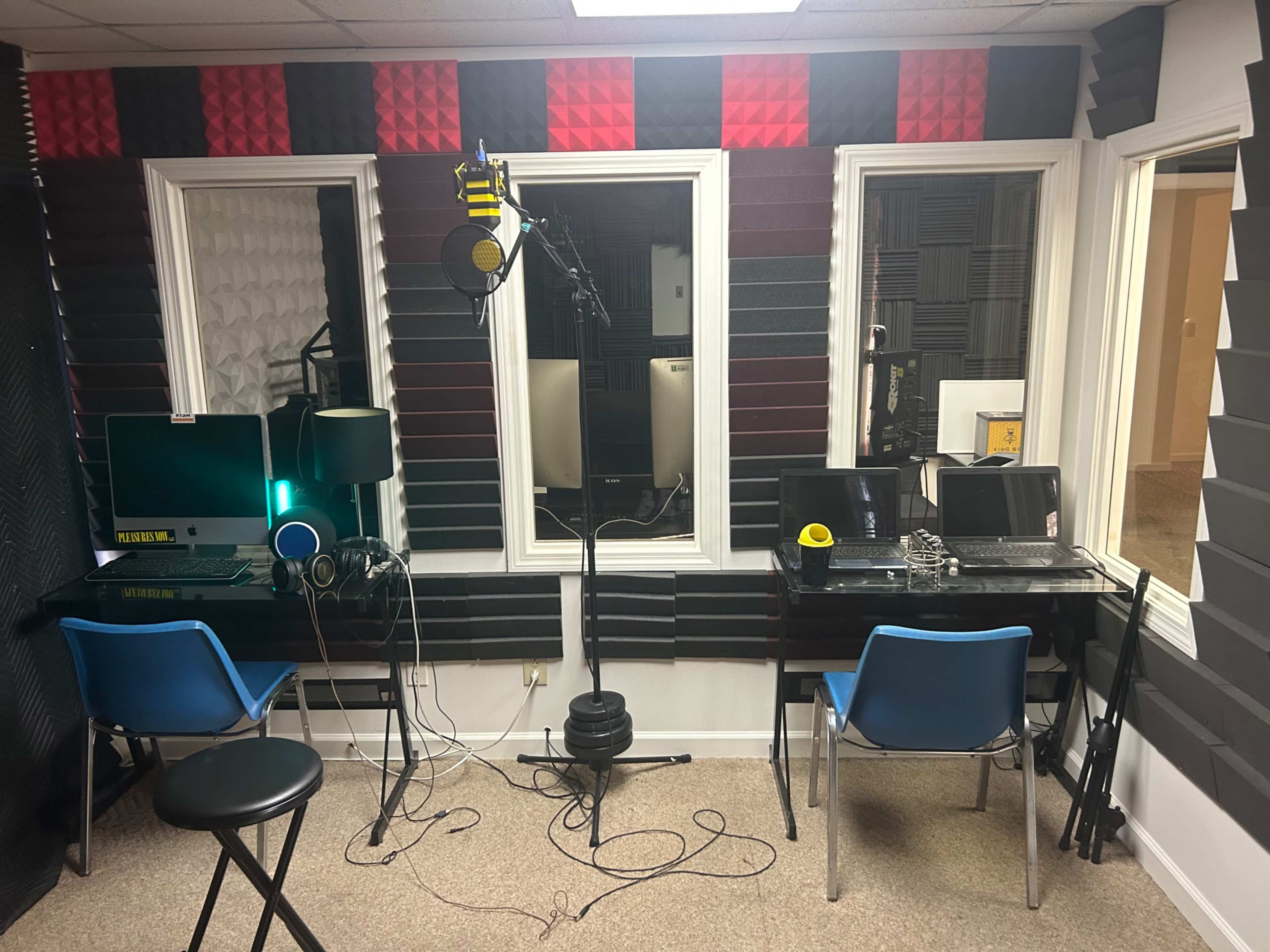 A sound studio with acoustic panels on the walls, two desk setups with laptops, a microphone on a stand, and chairs positioned around the space.