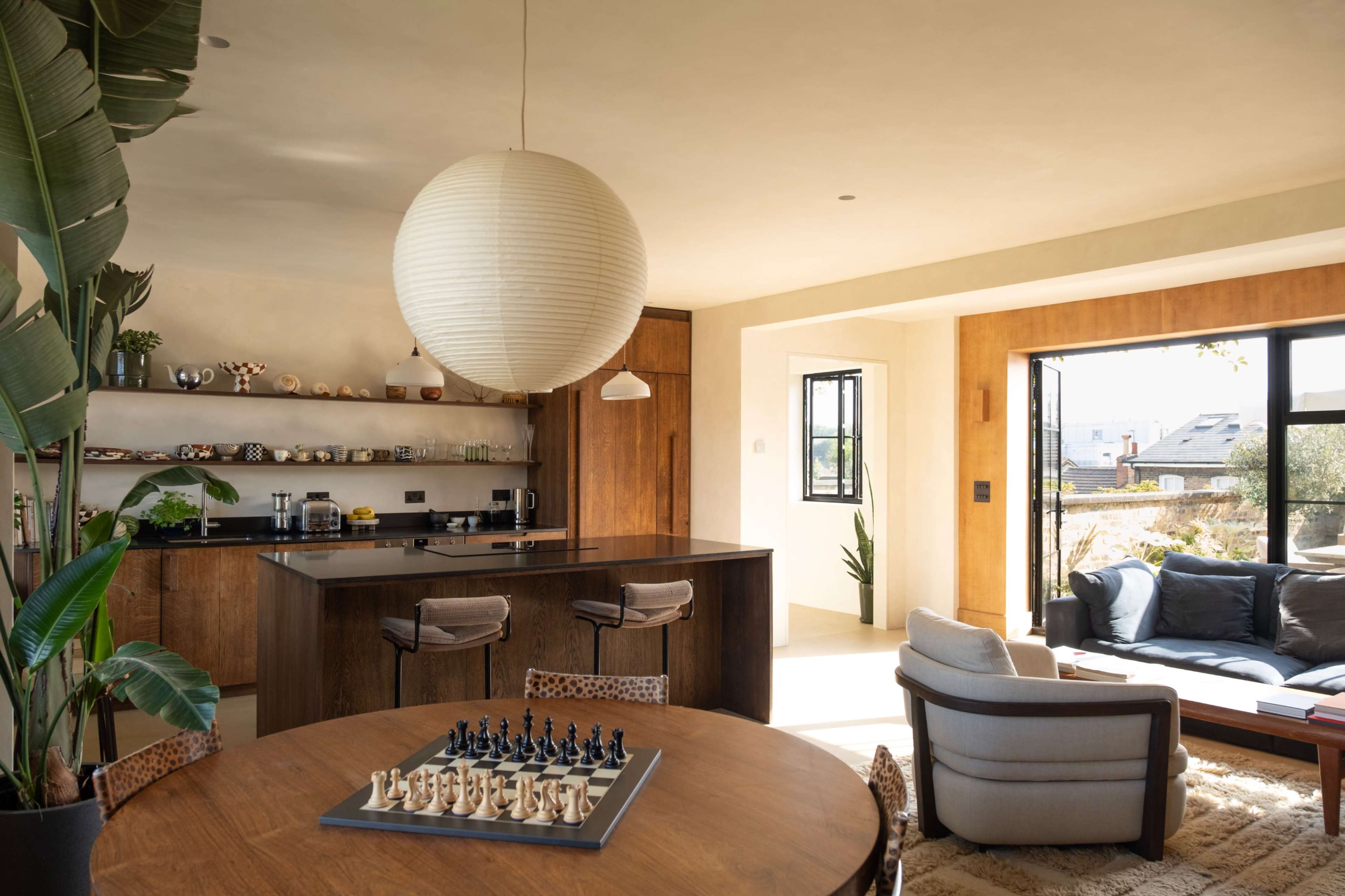 The image shows a modern interior space featuring a dining area with a round table, a chess set, and a kitchen bar with stools, alongside a living area with a sofa and large windows.