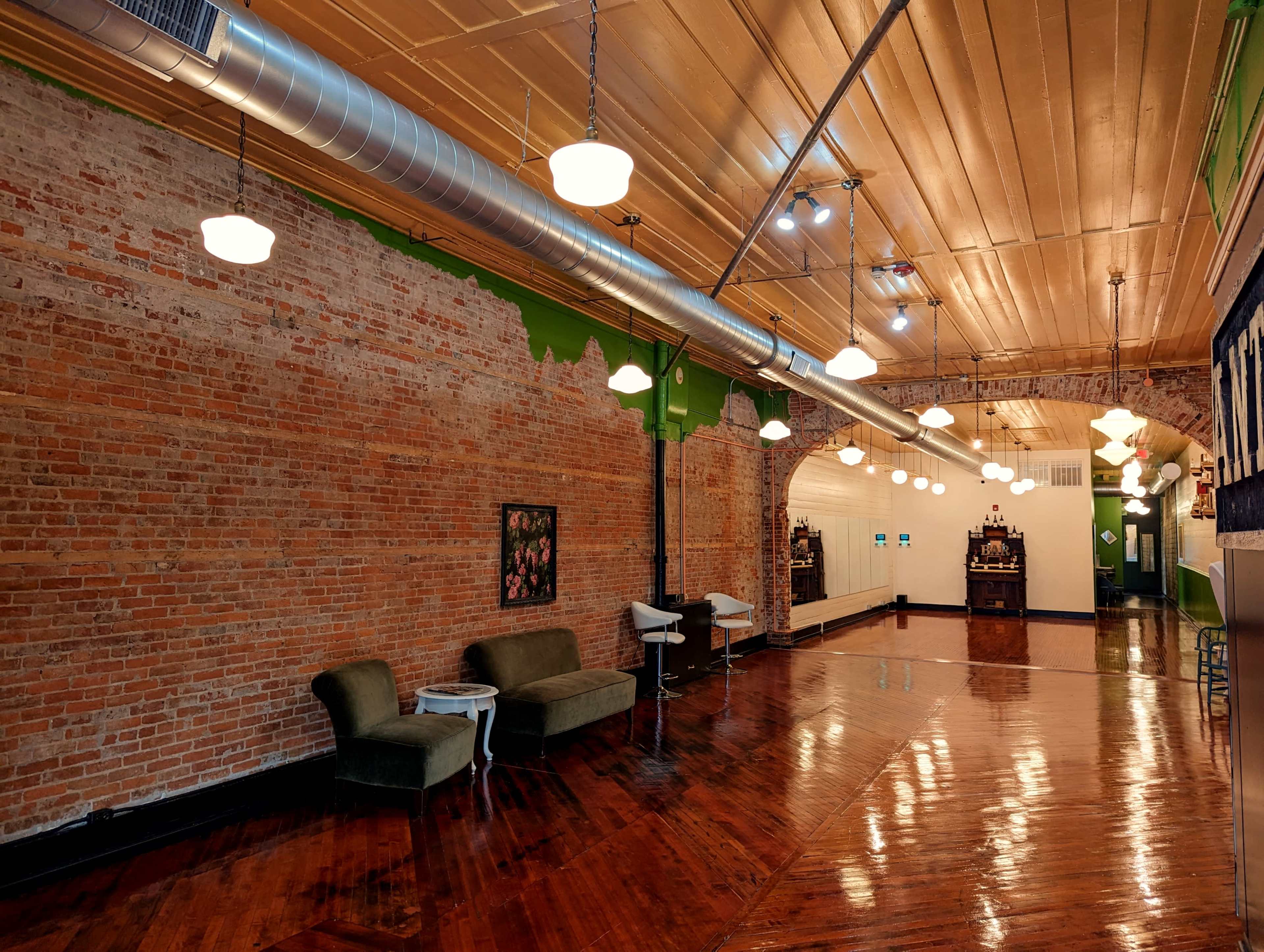 A long, narrow interior space with exposed brick walls, wooden flooring, and overhead lighting, featuring seating areas on one side.
