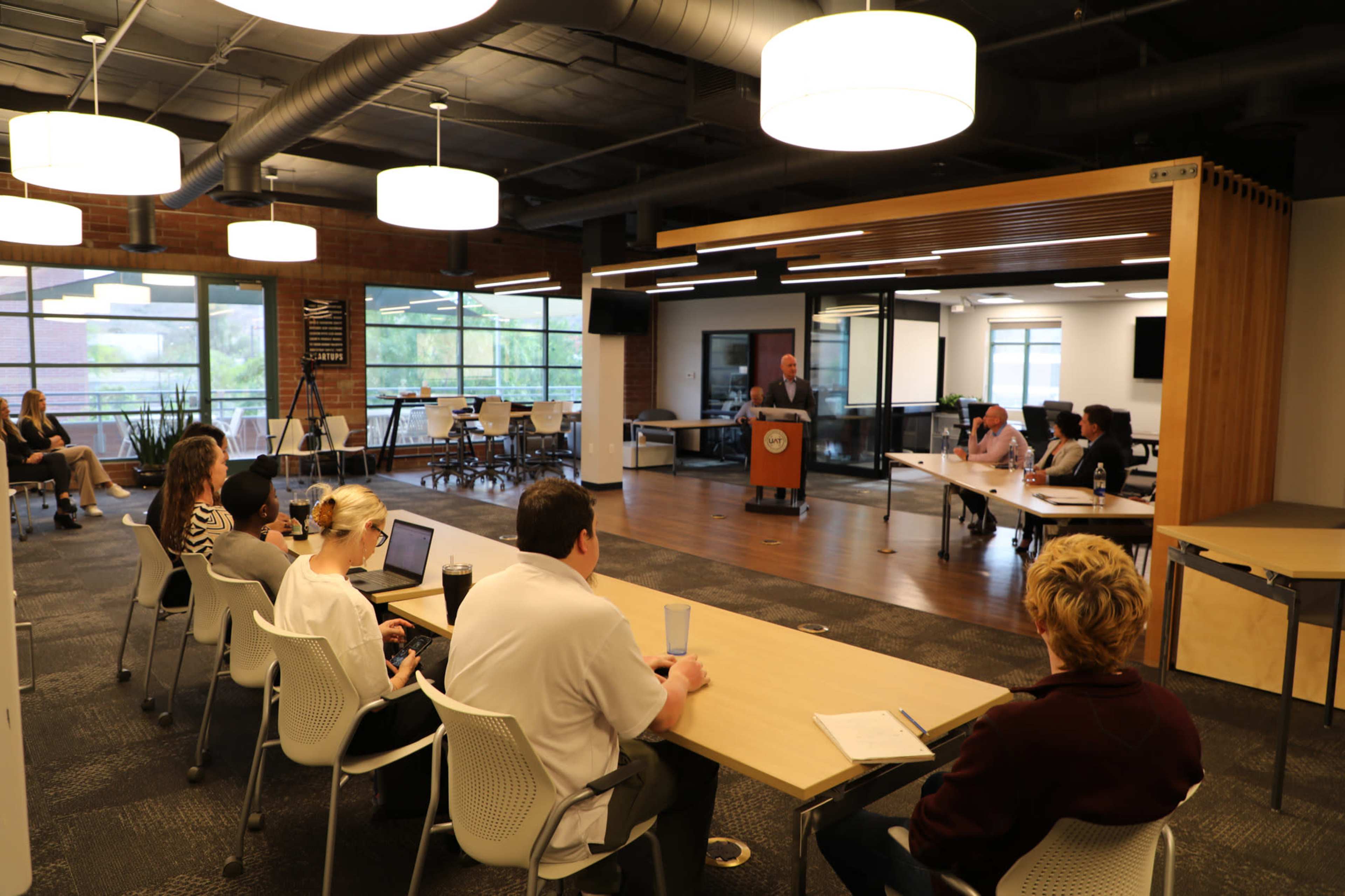 A group of people sits at tables in a conference room while a speaker presents at a podium in front of them.