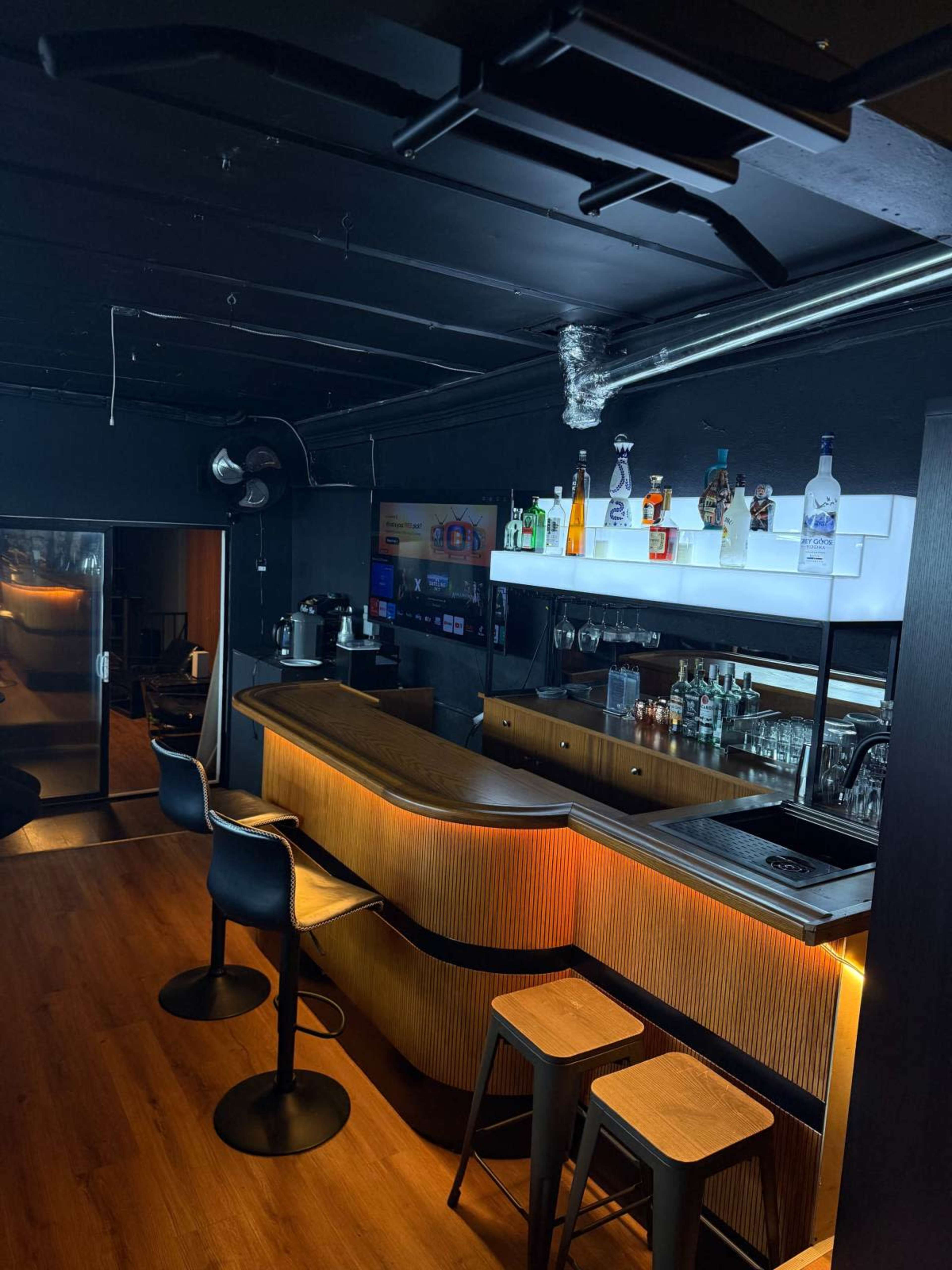 The image shows a modern bar area with a sleek wooden countertop, illuminated shelves displaying various bottles, and a mix of bar stools and wooden seating.