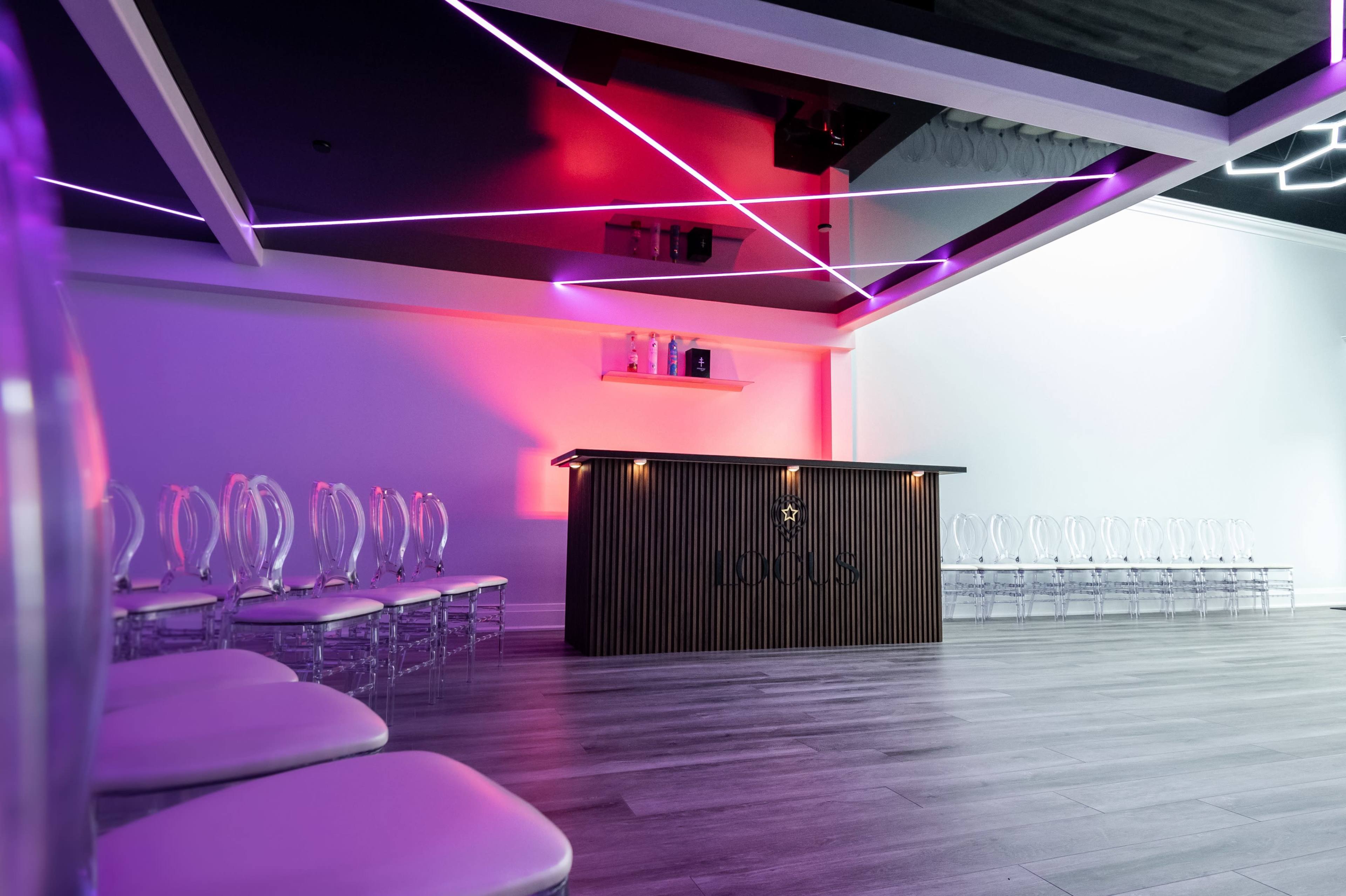 A modern event space features a wooden bar with illuminated shelves and rows of transparent chairs arranged on one side.