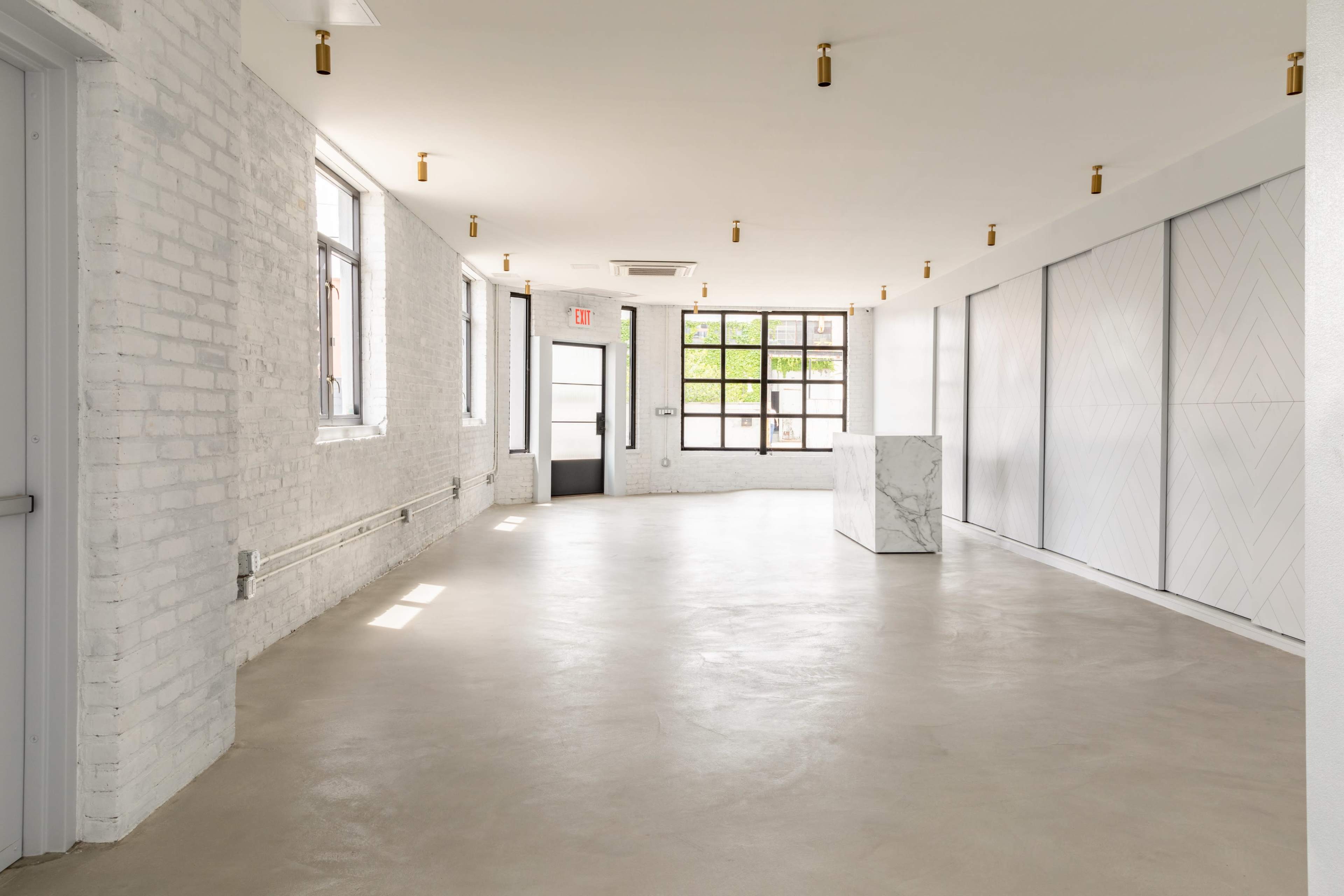 A spacious, well-lit room features white brick walls, concrete flooring, and large windows.