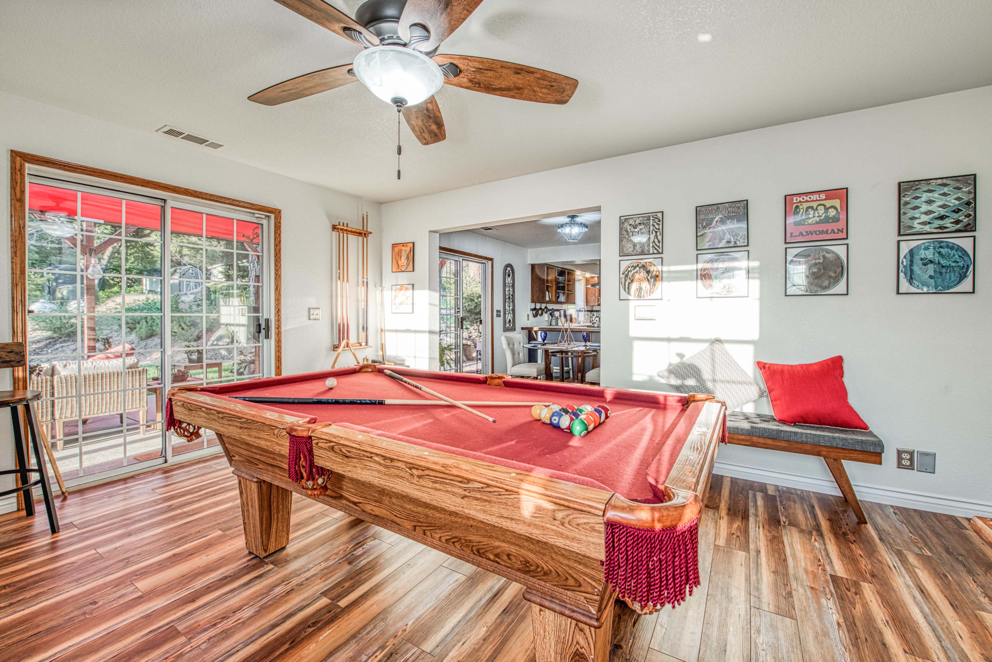A well-lit game room featuring a red felt pool table, wooden ceiling fan, and walls adorned with framed artwork.