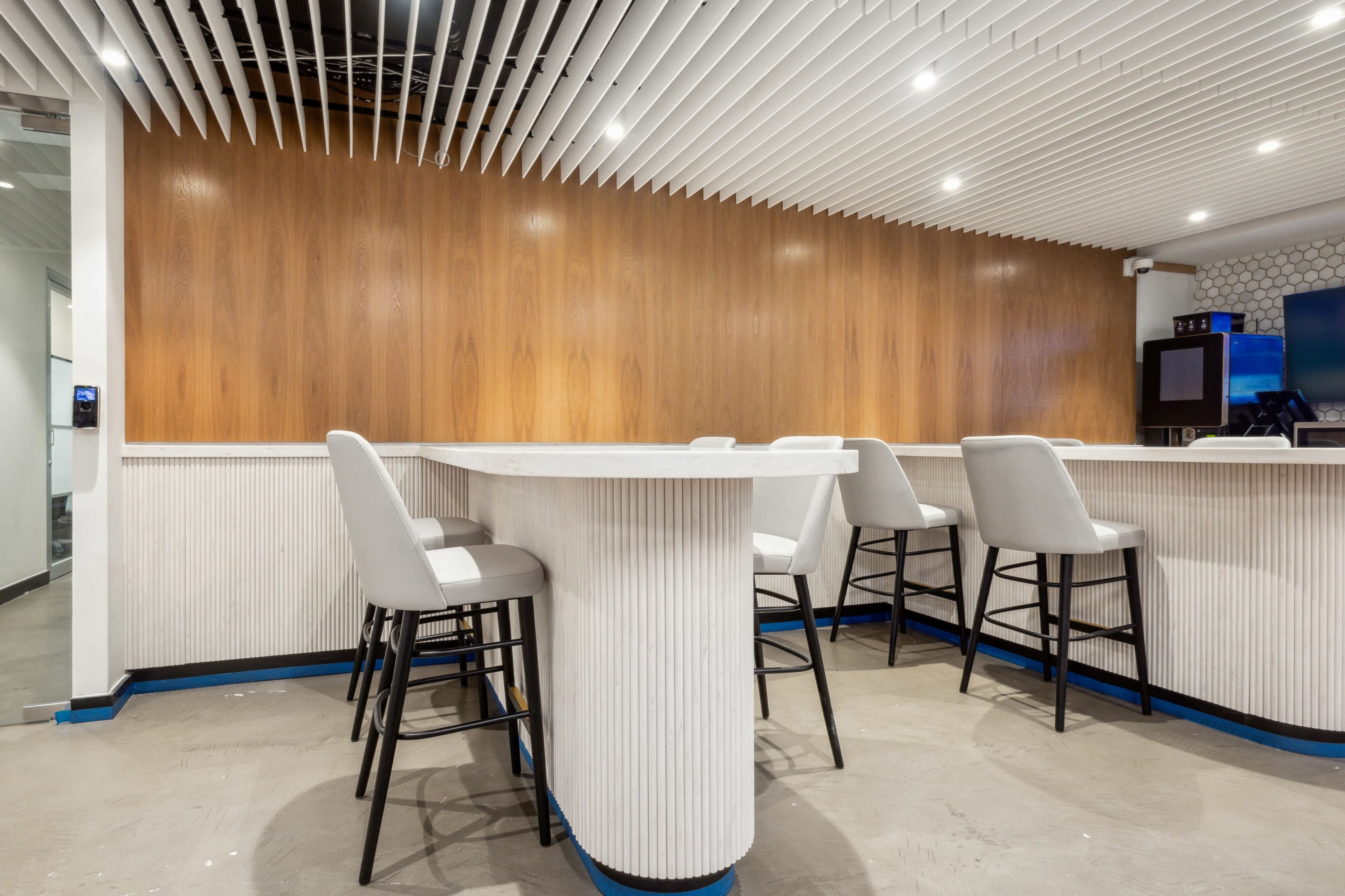 The image shows a modern bar area with a white counter and several black and white stools, set against a wooden-paneled wall and ceiling with linear designs.