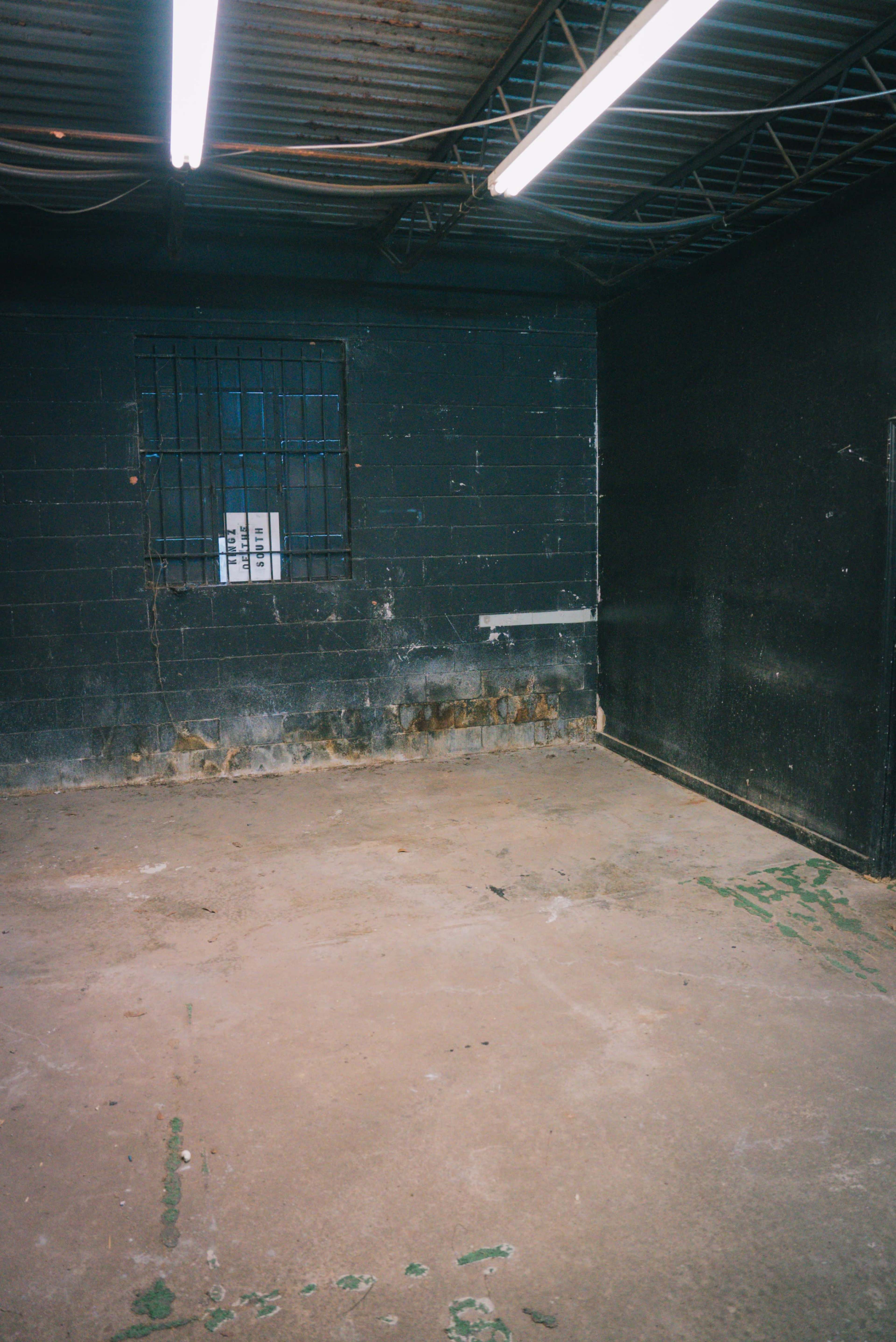 The image shows a sparsely furnished room with a concrete floor, black-painted walls, a barred window, and visible wear on the surface.