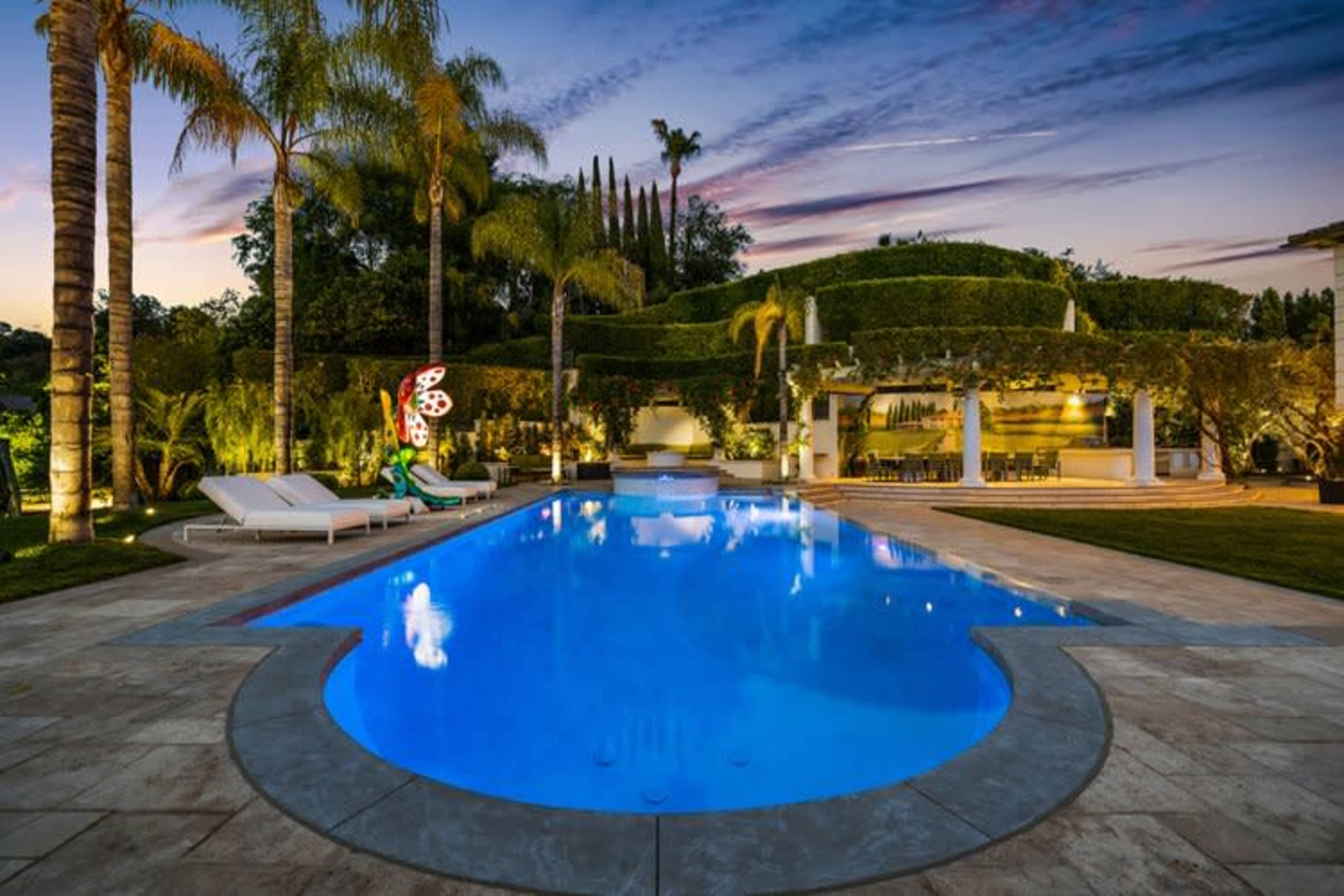 A brightly lit swimming pool surrounded by lush greenery and lounge chairs, with a vibrant sculpture and outdoor dining area in the background during sunset.