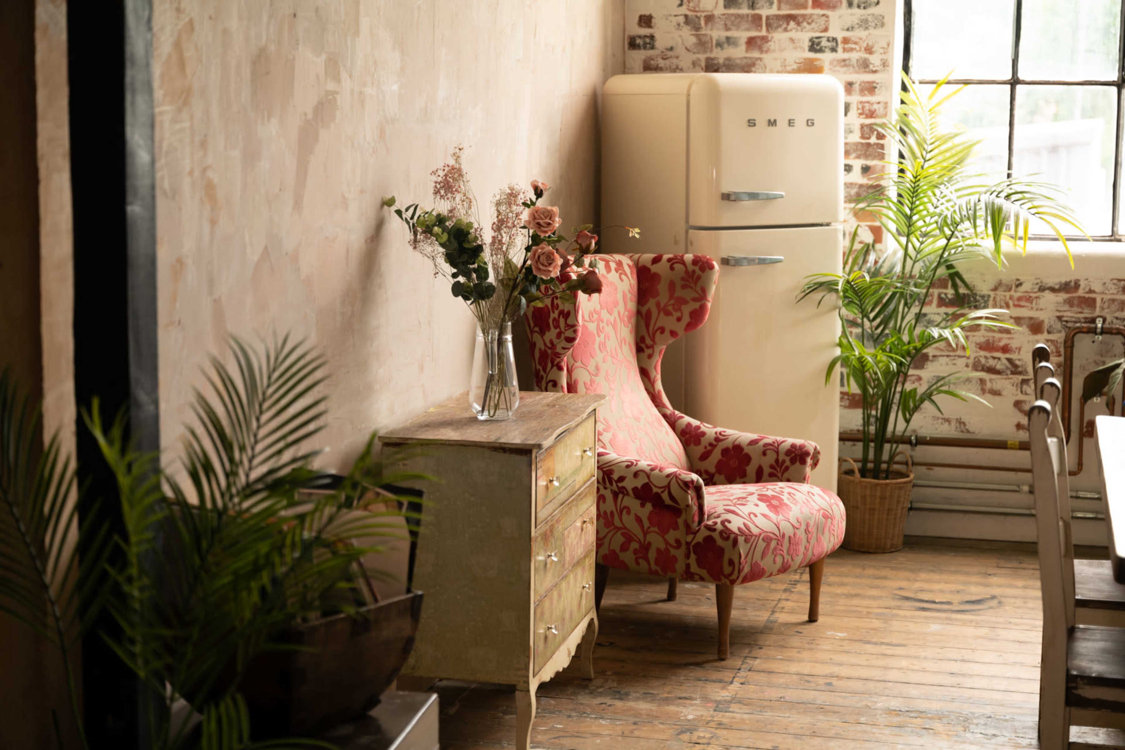 A cozy interior features a vintage Smeg refrigerator, a floral armchair, a wooden chest, and potted plants against a textured wall.