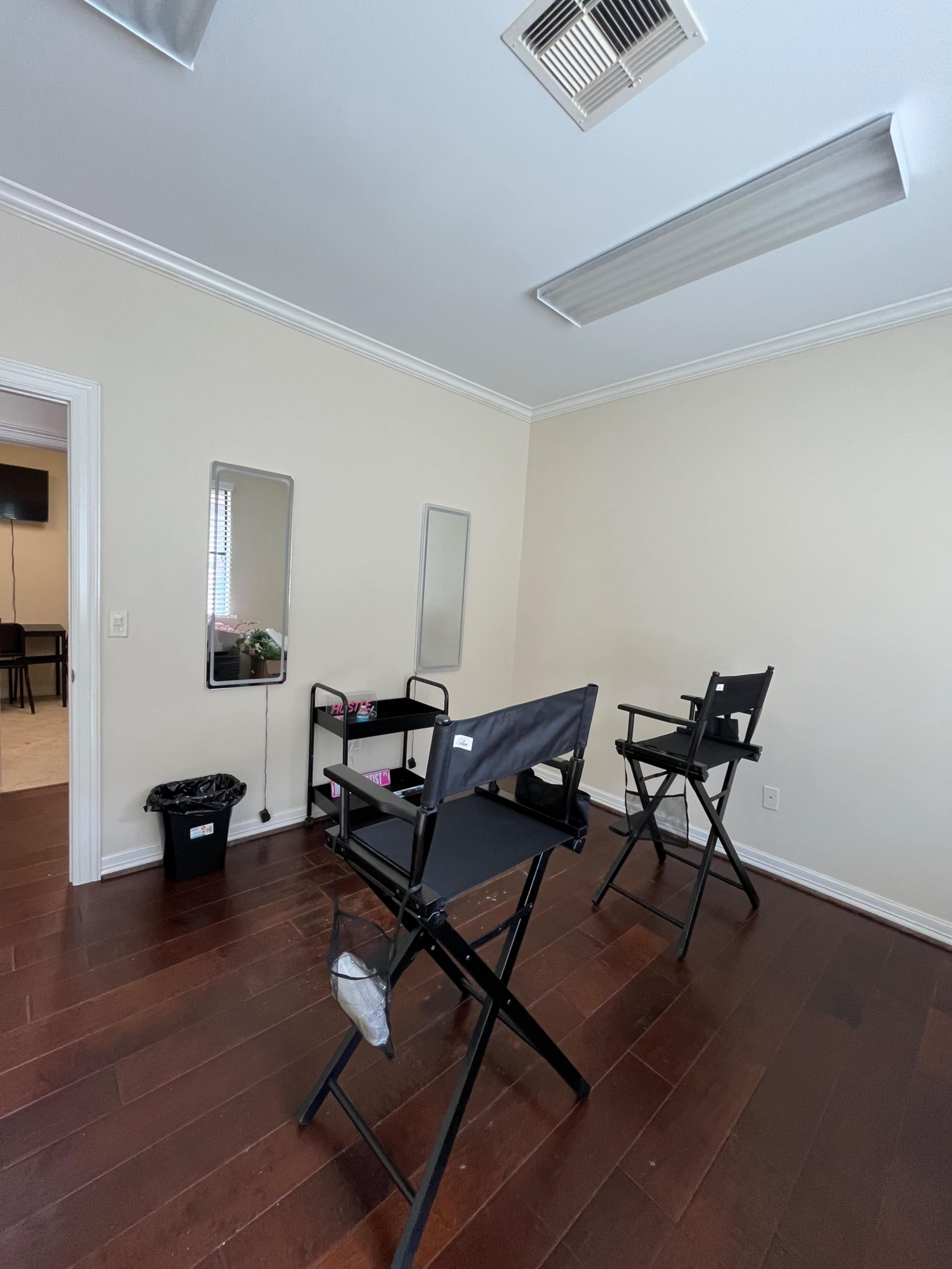 The image shows a small, well-lit room with two black director's chairs, a table with items on it, and a waste bin, all positioned on wooden flooring.