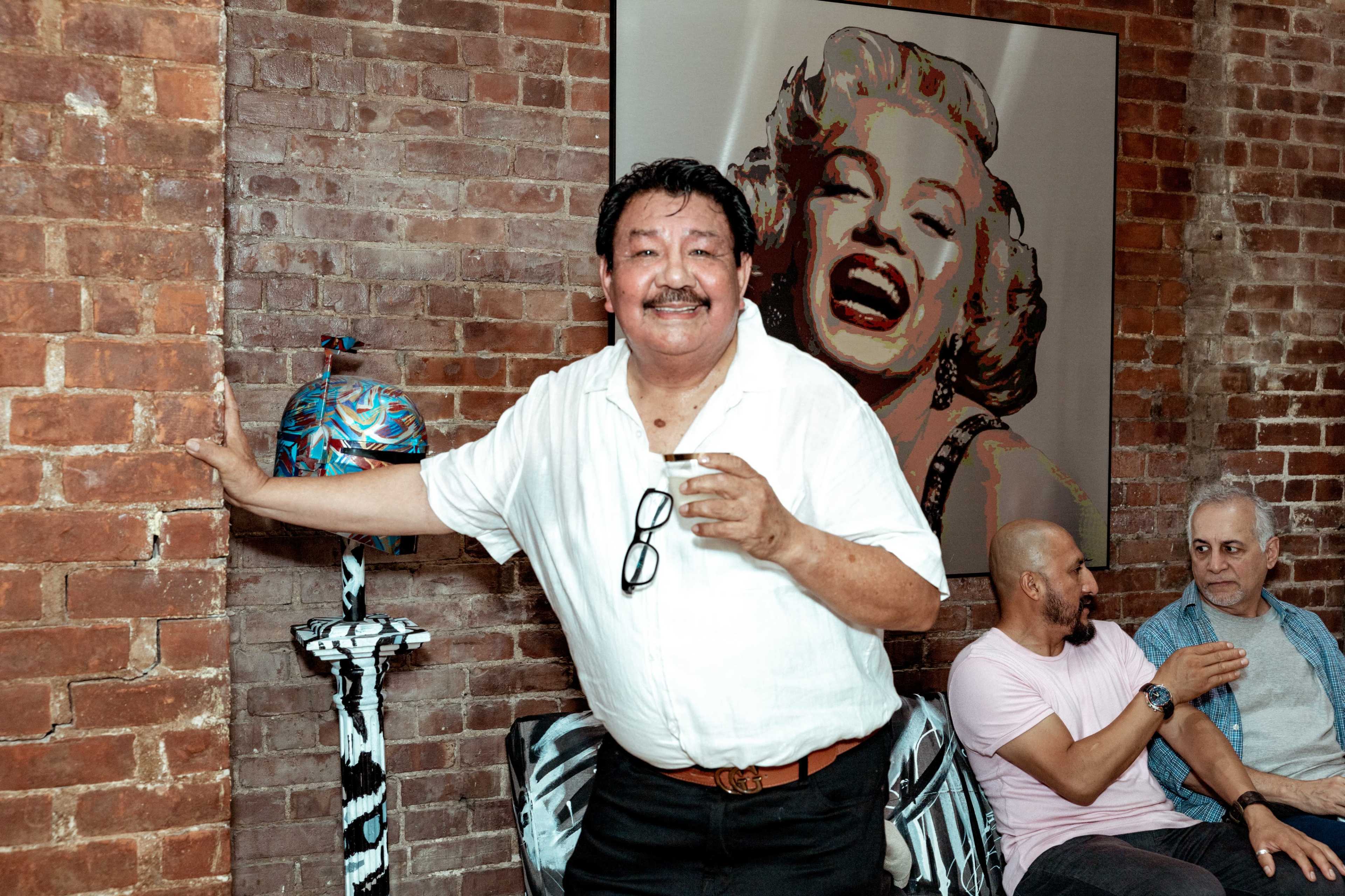 A man stands near a brick wall holding a drink, with a large portrait of Marilyn Monroe in the background, while two other men chat on a couch.