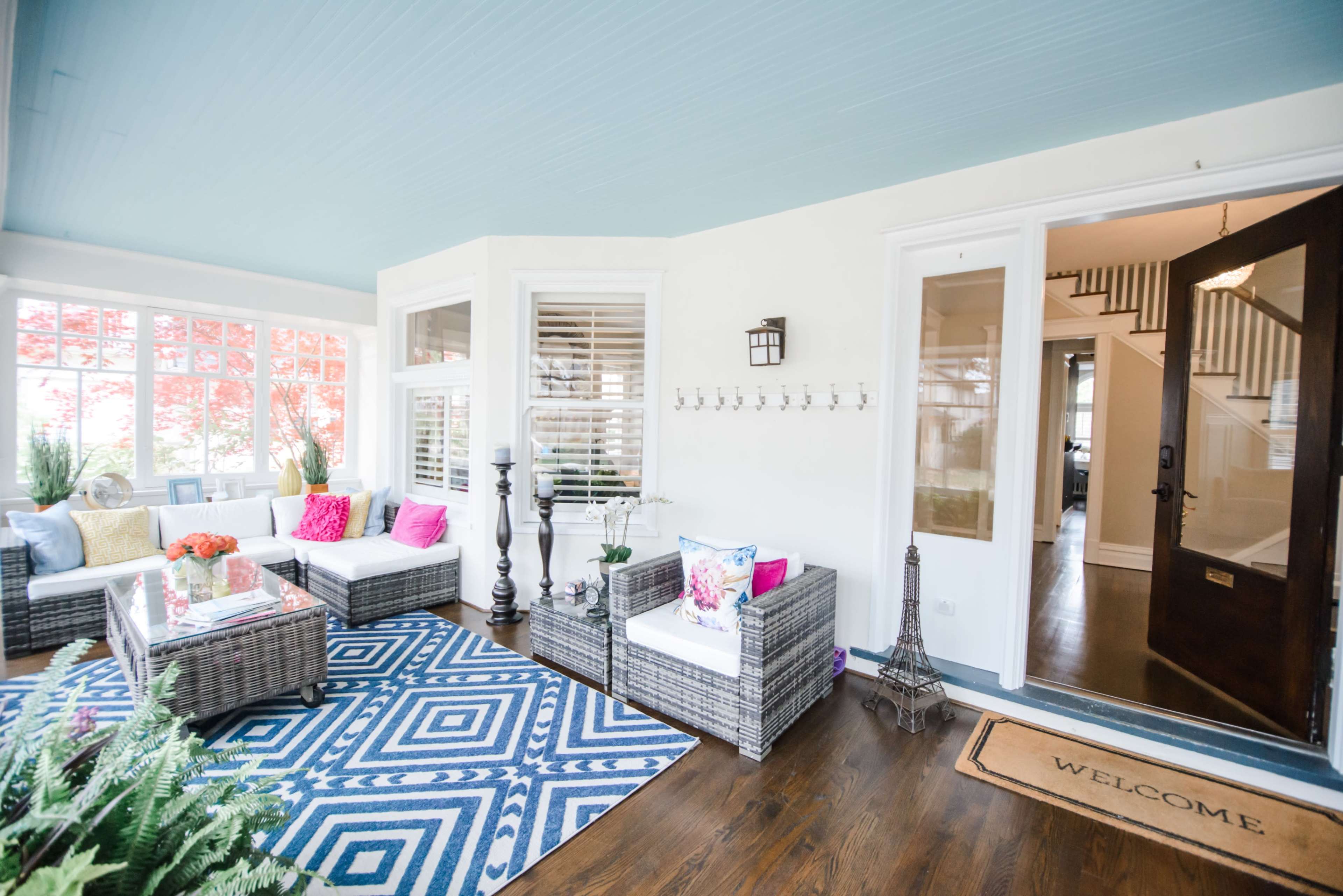 A welcoming sunroom with wicker furniture, colorful cushions, and a large entrance leading to a staircase.