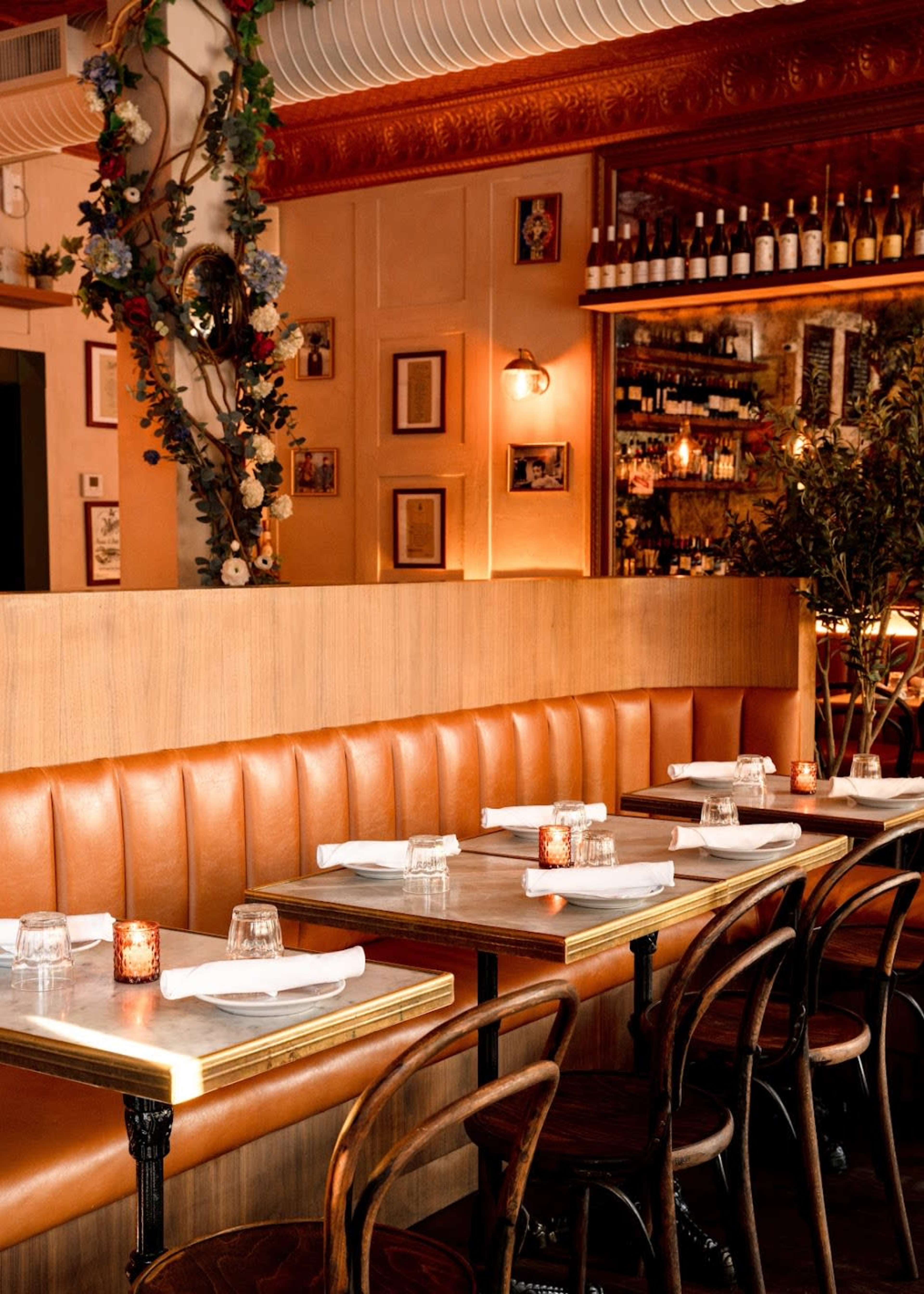 The image shows a cozy restaurant interior with brown leather banquettes, wooden tables, and a well-stocked wine shelf in the background.