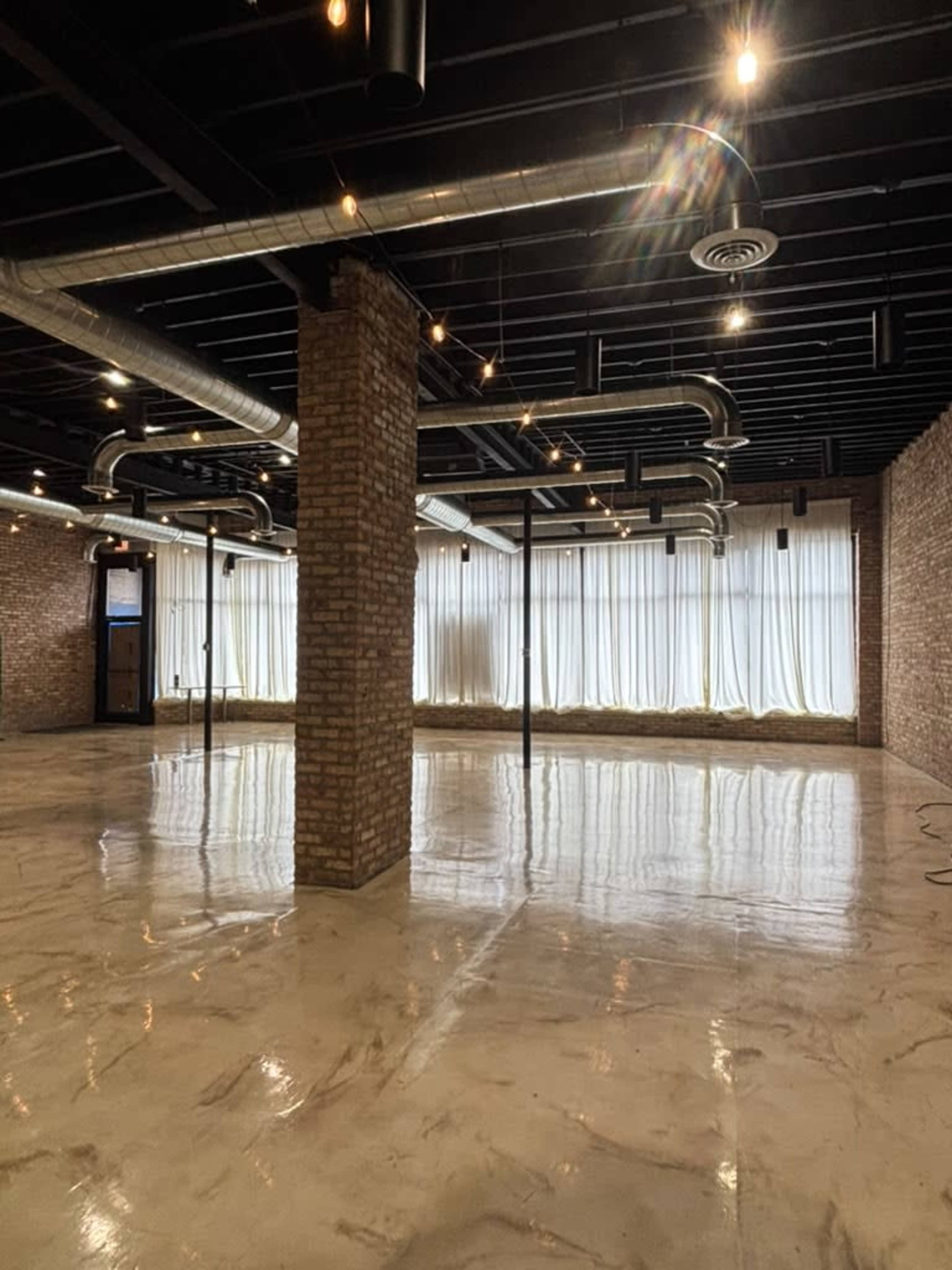 The image shows a spacious, unfinished interior with exposed brick walls, polished concrete floors, and visible ductwork on the ceiling.