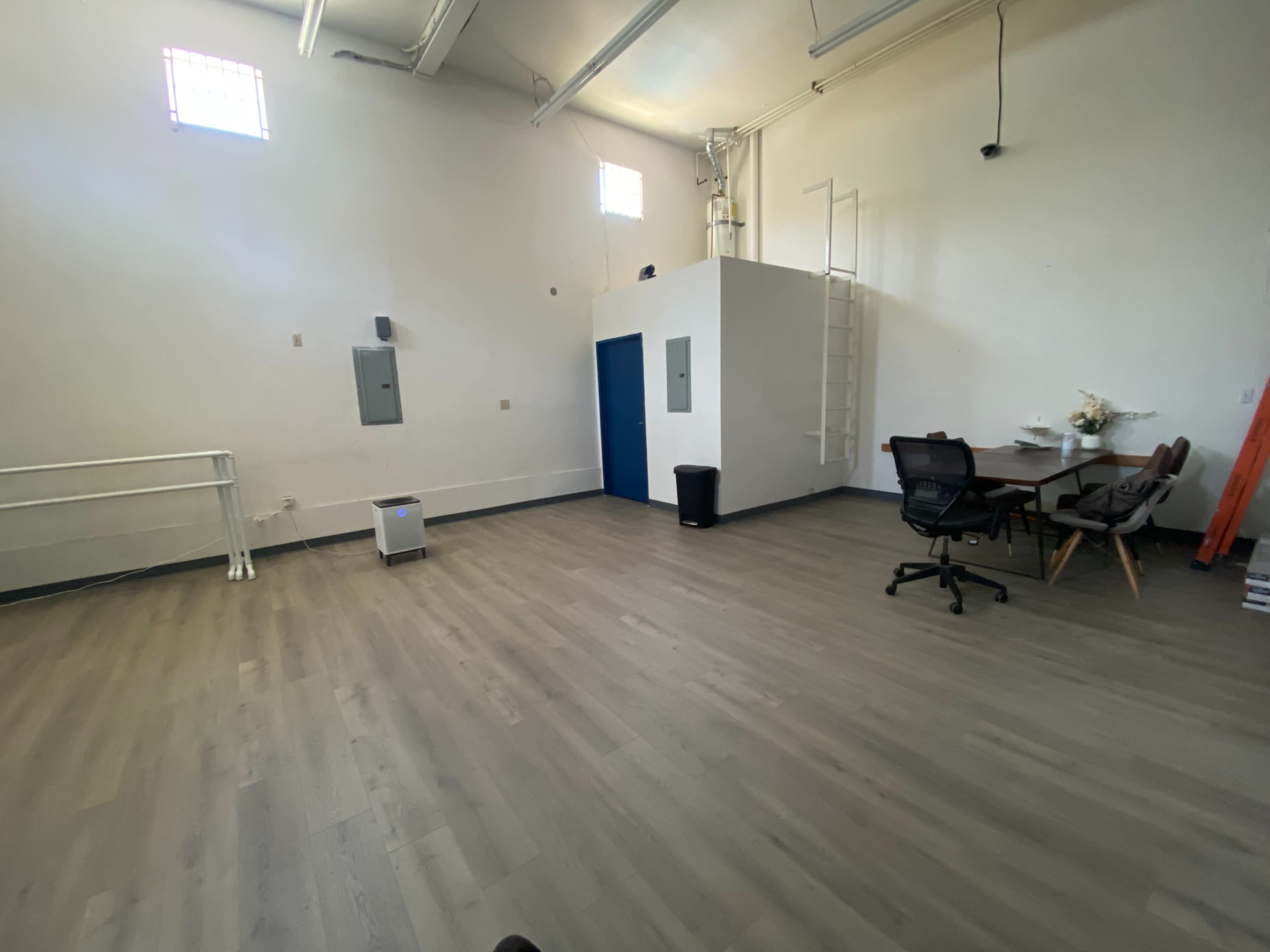 An empty room with light-colored flooring, a white wall-mounted storage unit, a small table with chairs, and a ladder leading to a loft area.