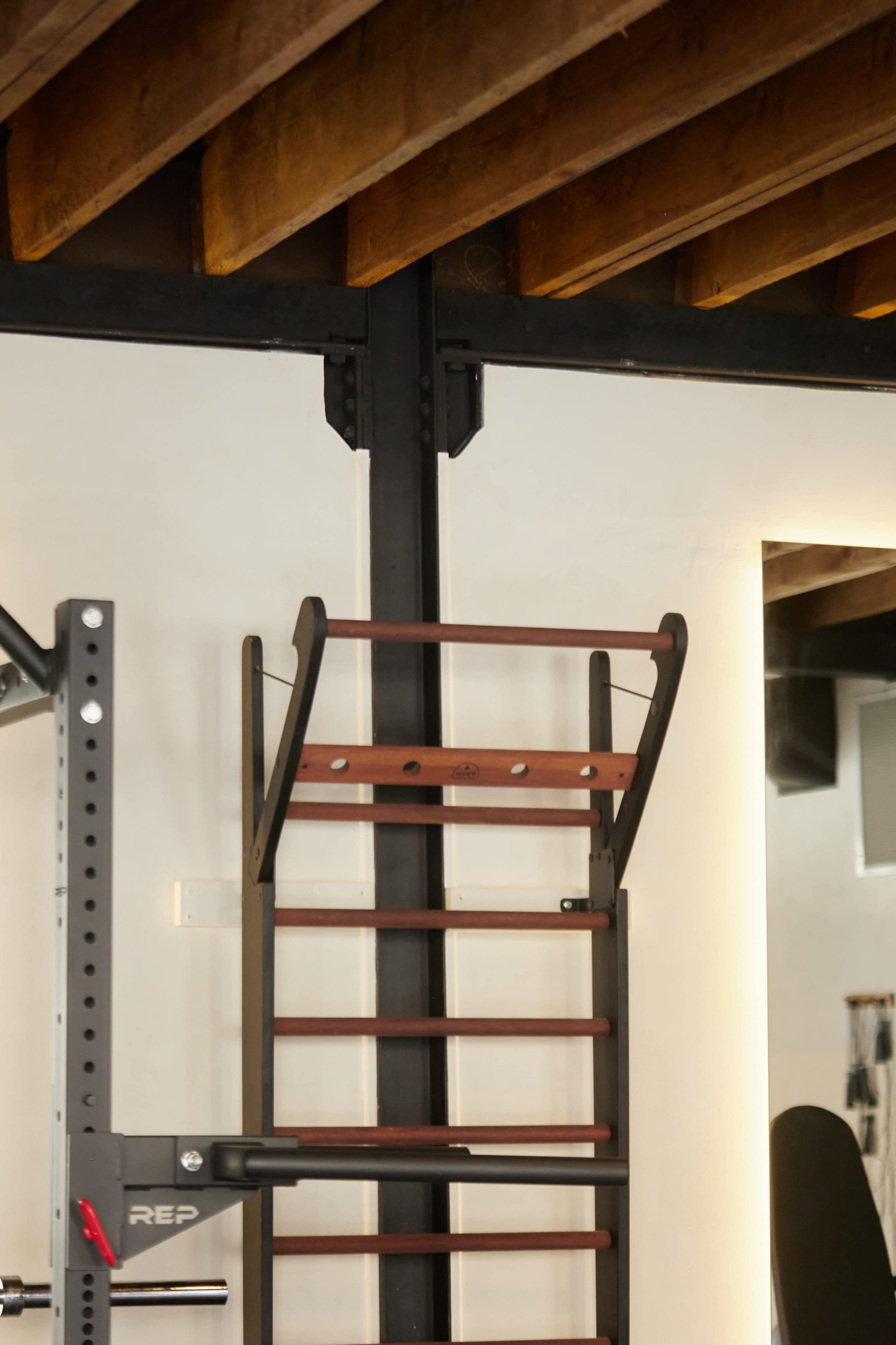 A wooden gym ladder mounted against a wall in a fitness space with exposed beams overhead.