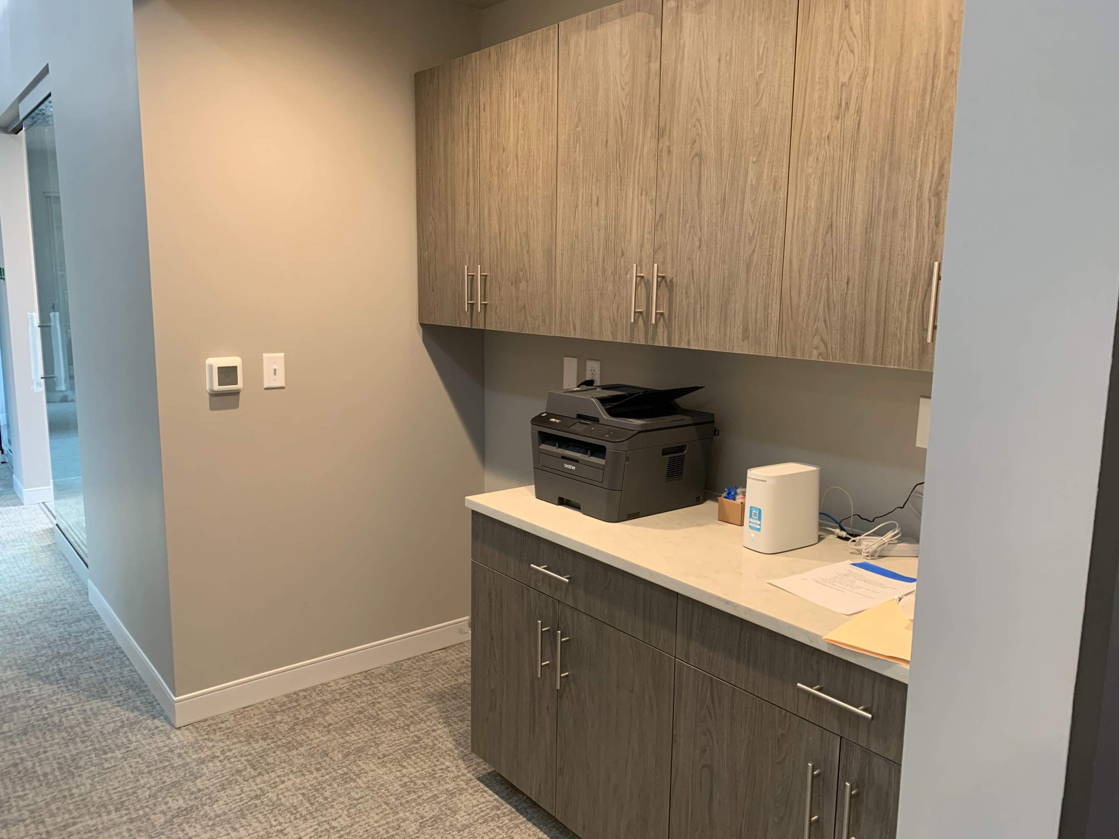 A light-colored office corner with gray cabinets, a printer on the countertop, and documents on the surface.