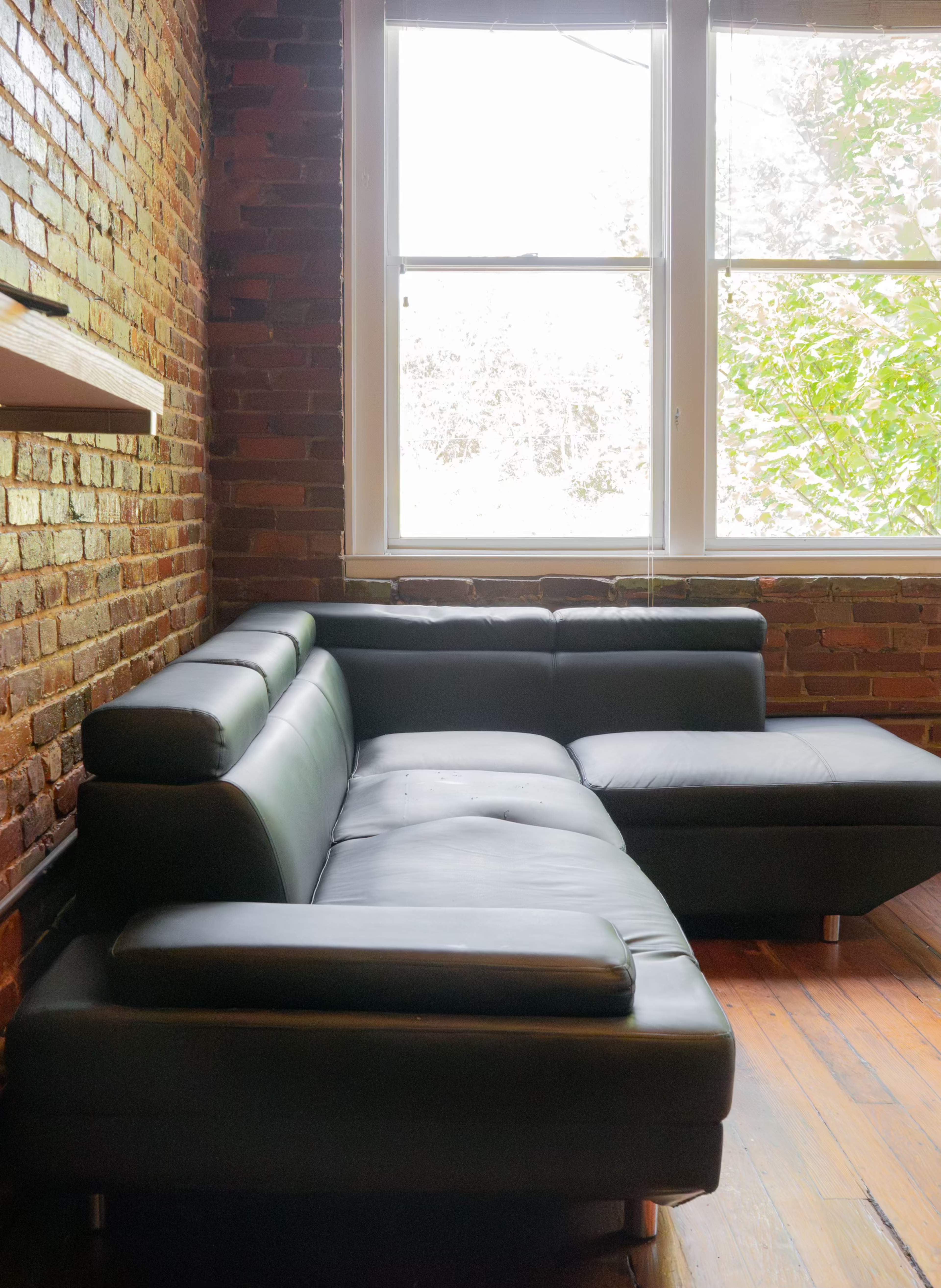 A black leather sectional sofa is positioned against a brick wall near two large windows.