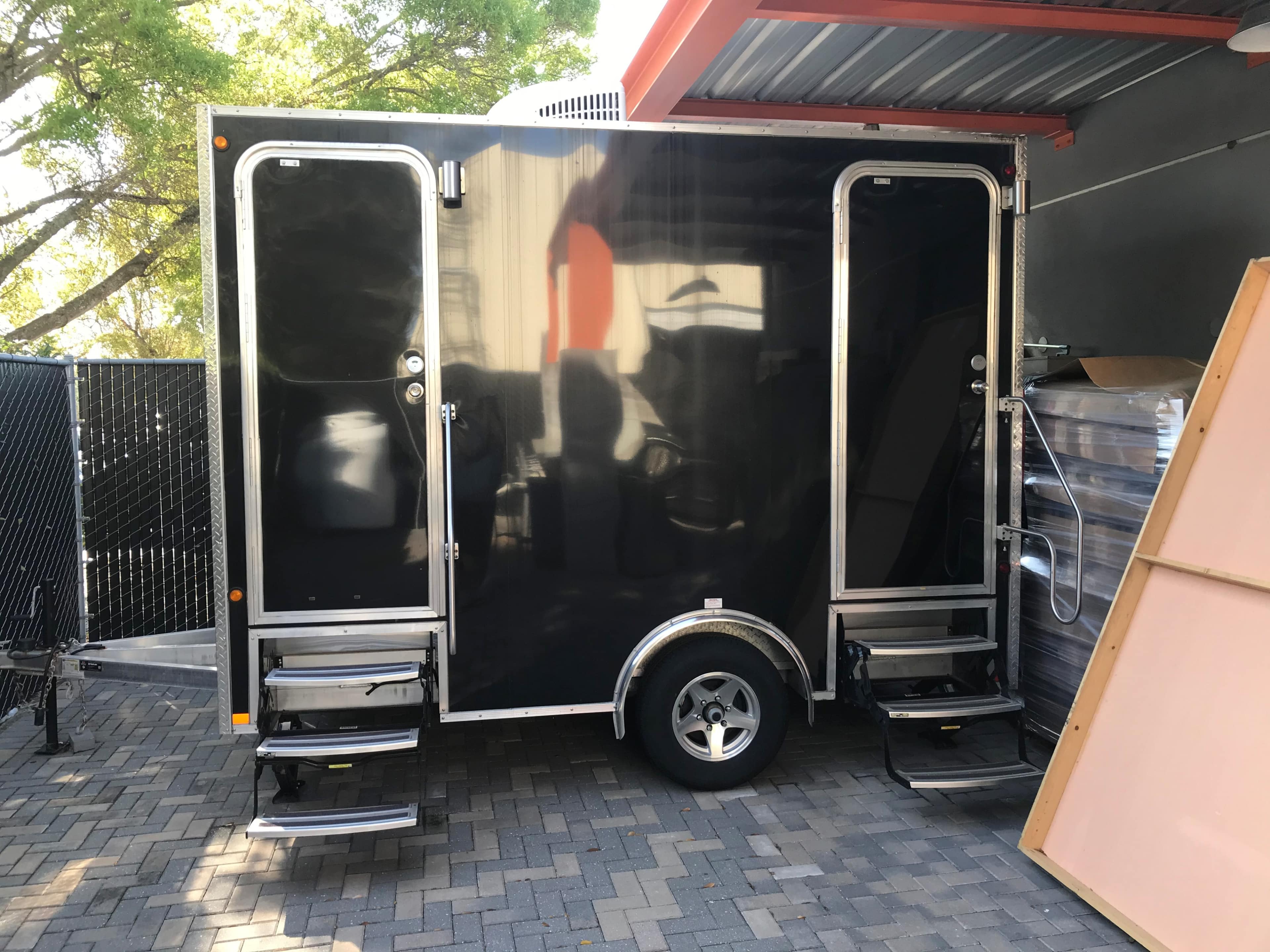 A black trailer with dual entry doors and steps, parked on patterned paving stones near a building.