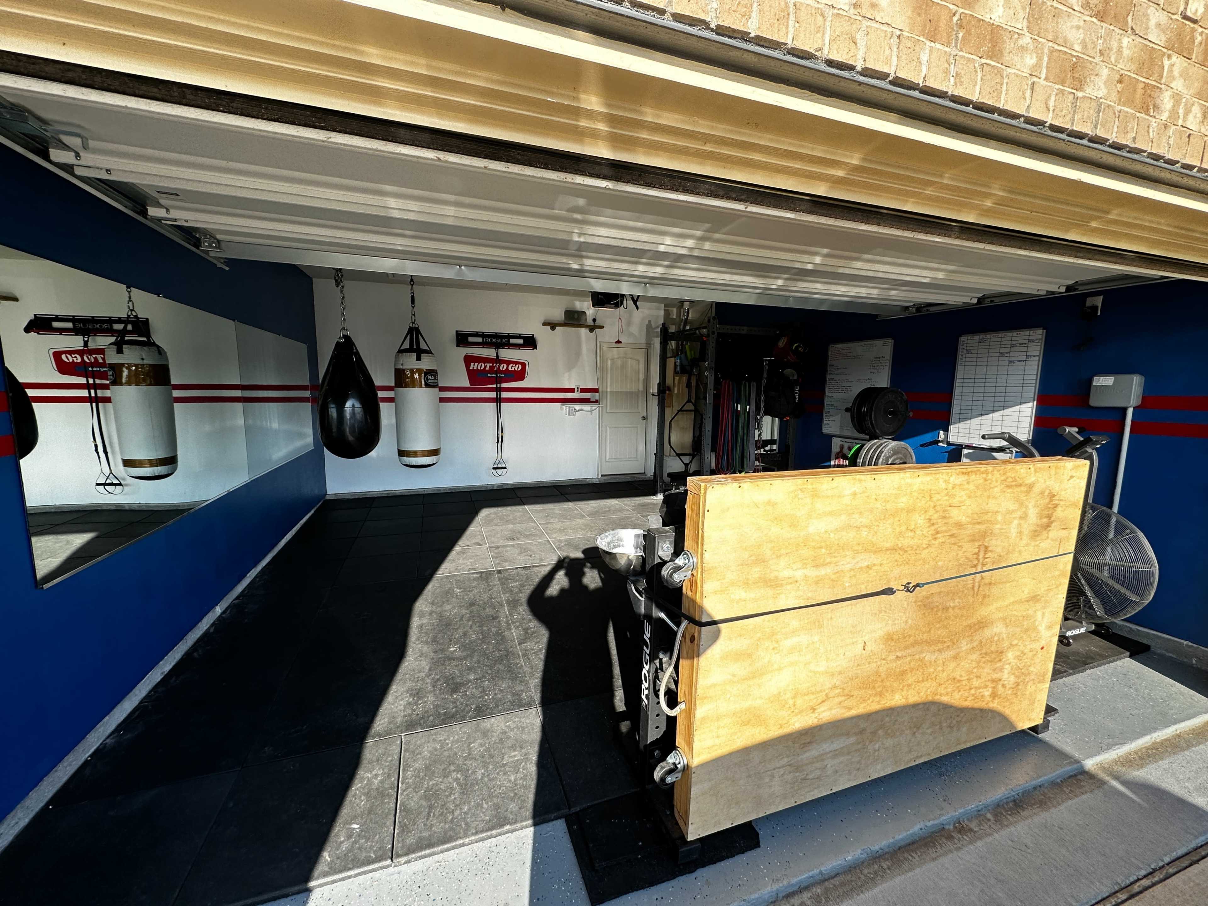 A home gym in a garage with punching bags, a weightlifting station, and a large mirror on the blue and white walls.