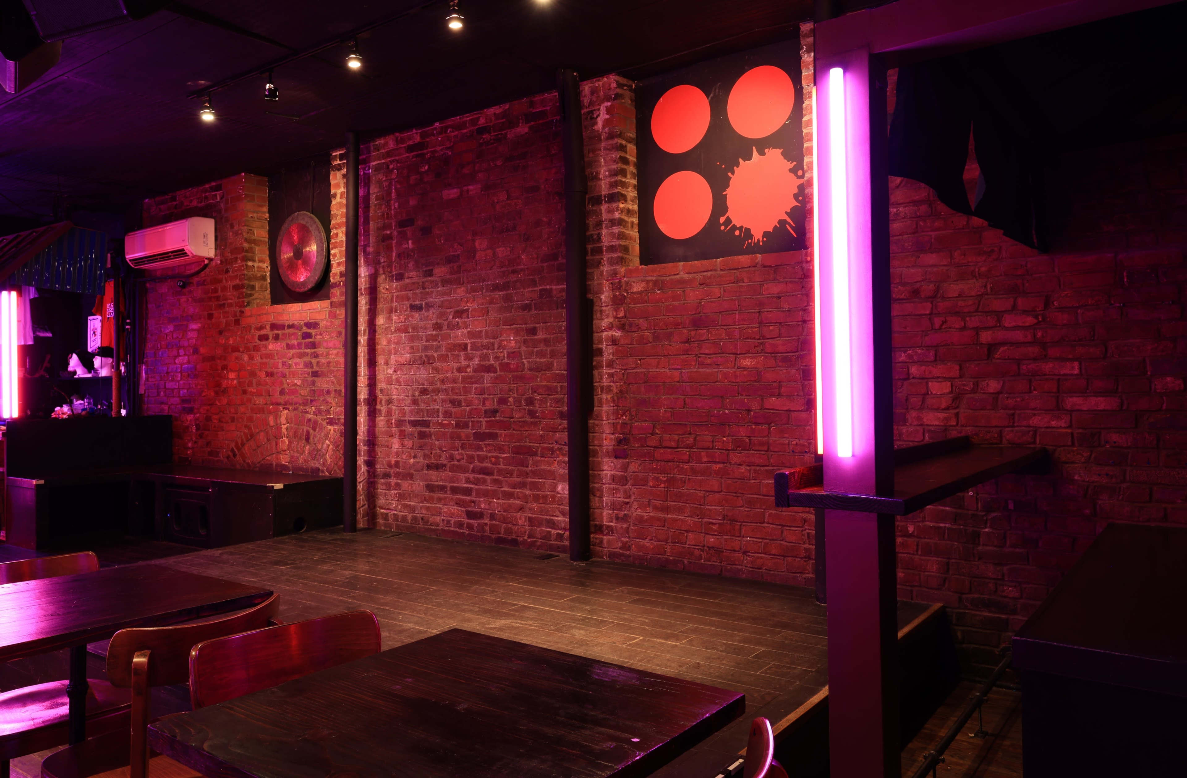 The image shows an empty brick-walled room with a small stage, colorful artwork on the walls, and dim lighting.
