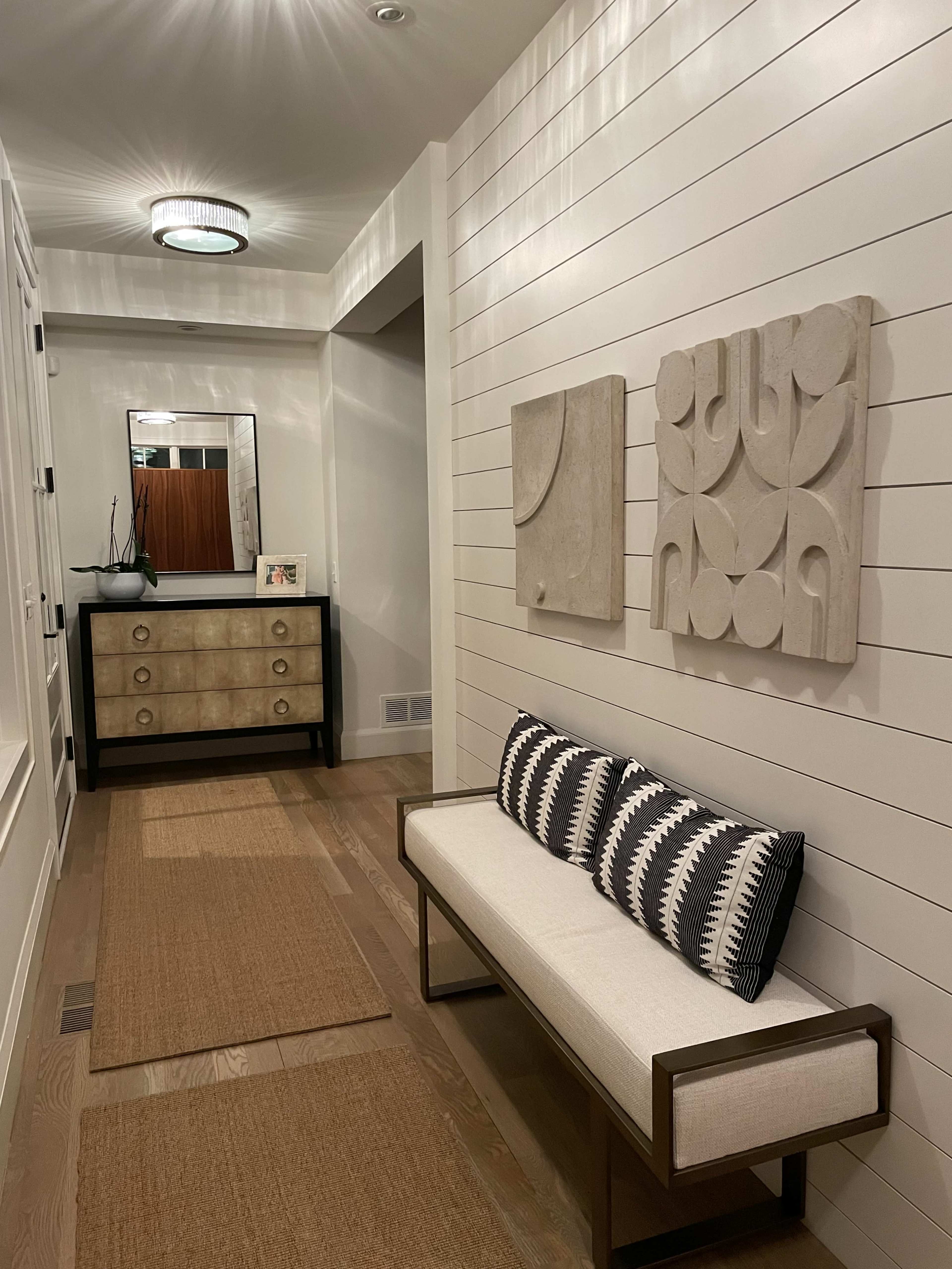 A modern hallway with a wooden floor, a bench with patterned pillows, a storage cabinet, and decorative wall art.
