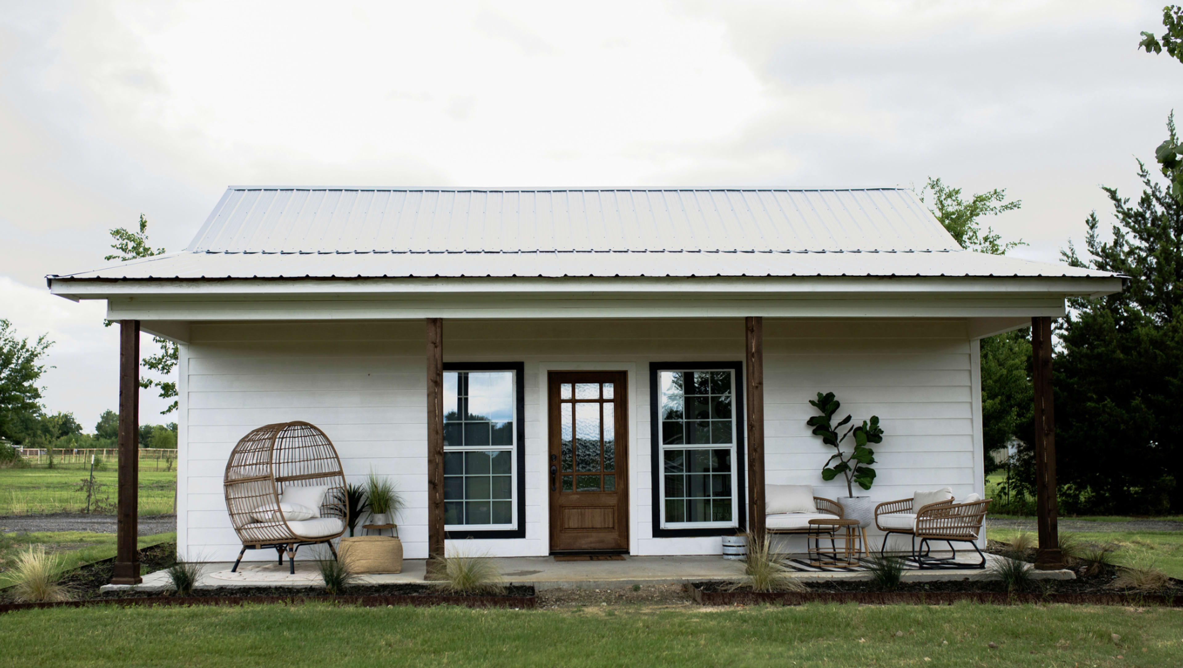 A white house with a metal roof, featuring a front porch with two sitting areas and large windows.