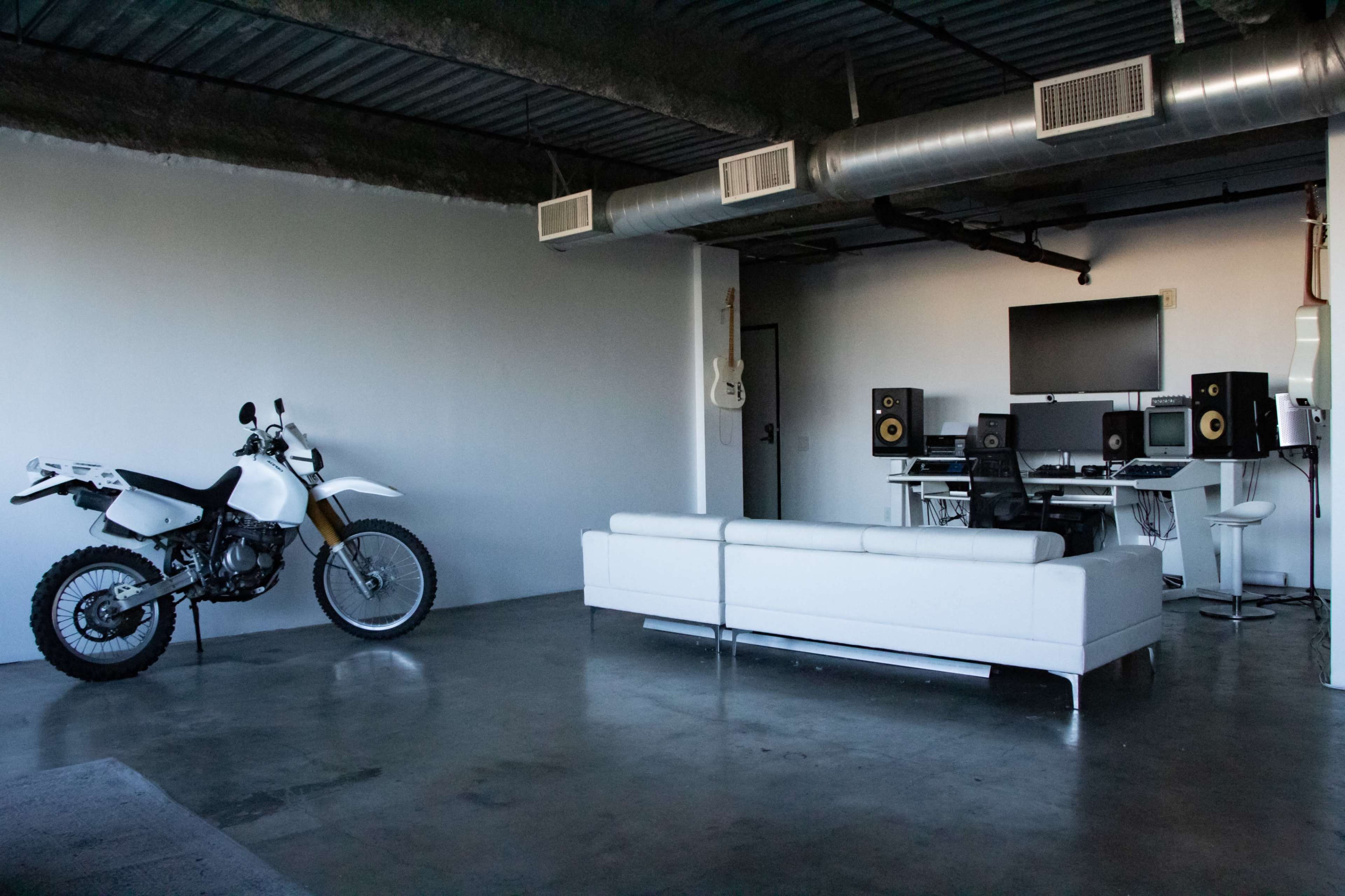 A white dirt bike is parked beside a modern living area featuring a white sofa and a music studio setup with speakers and a computer.