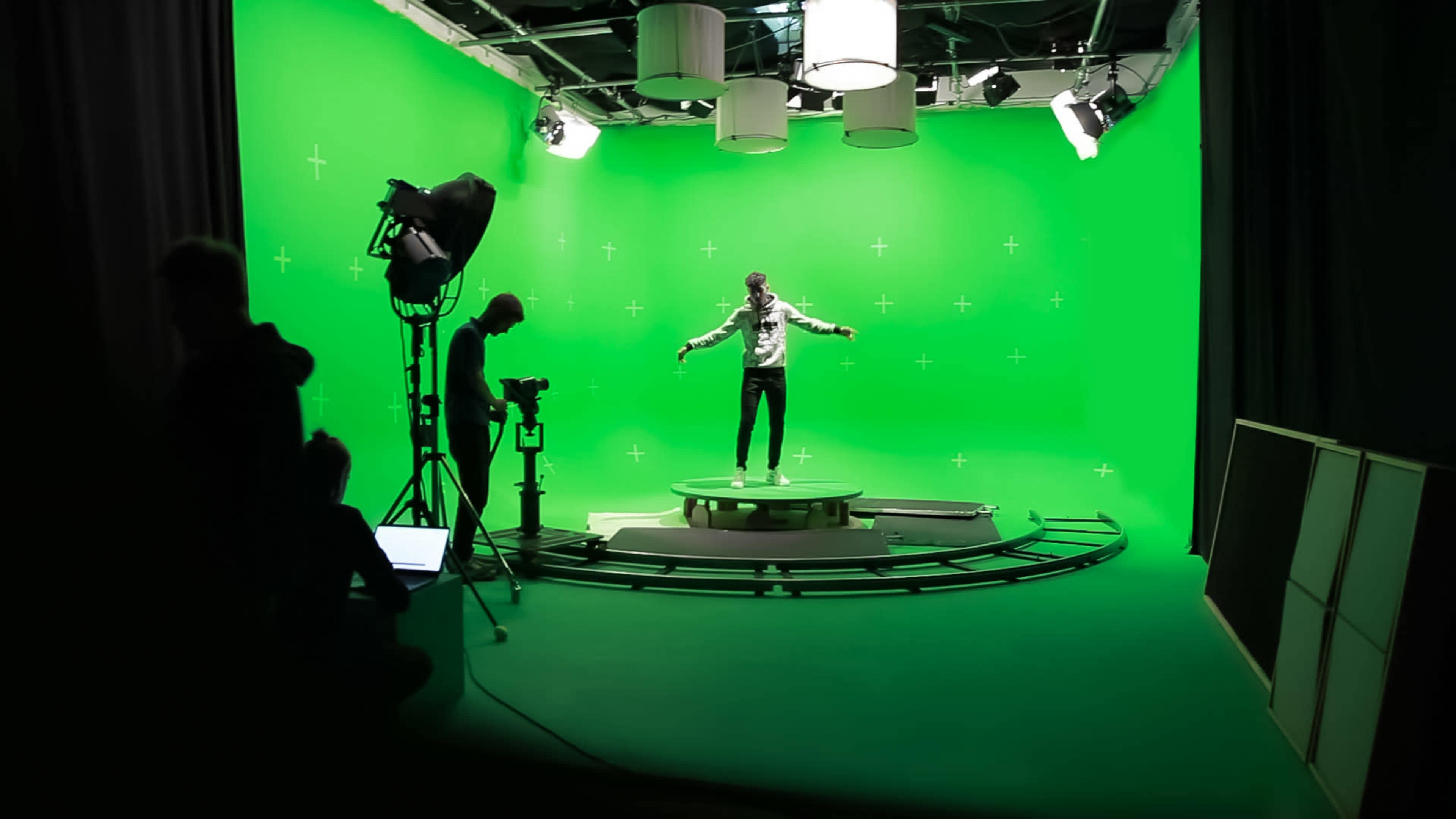 A person stands in the center of a green screen studio while crew members operate equipment in the foreground.