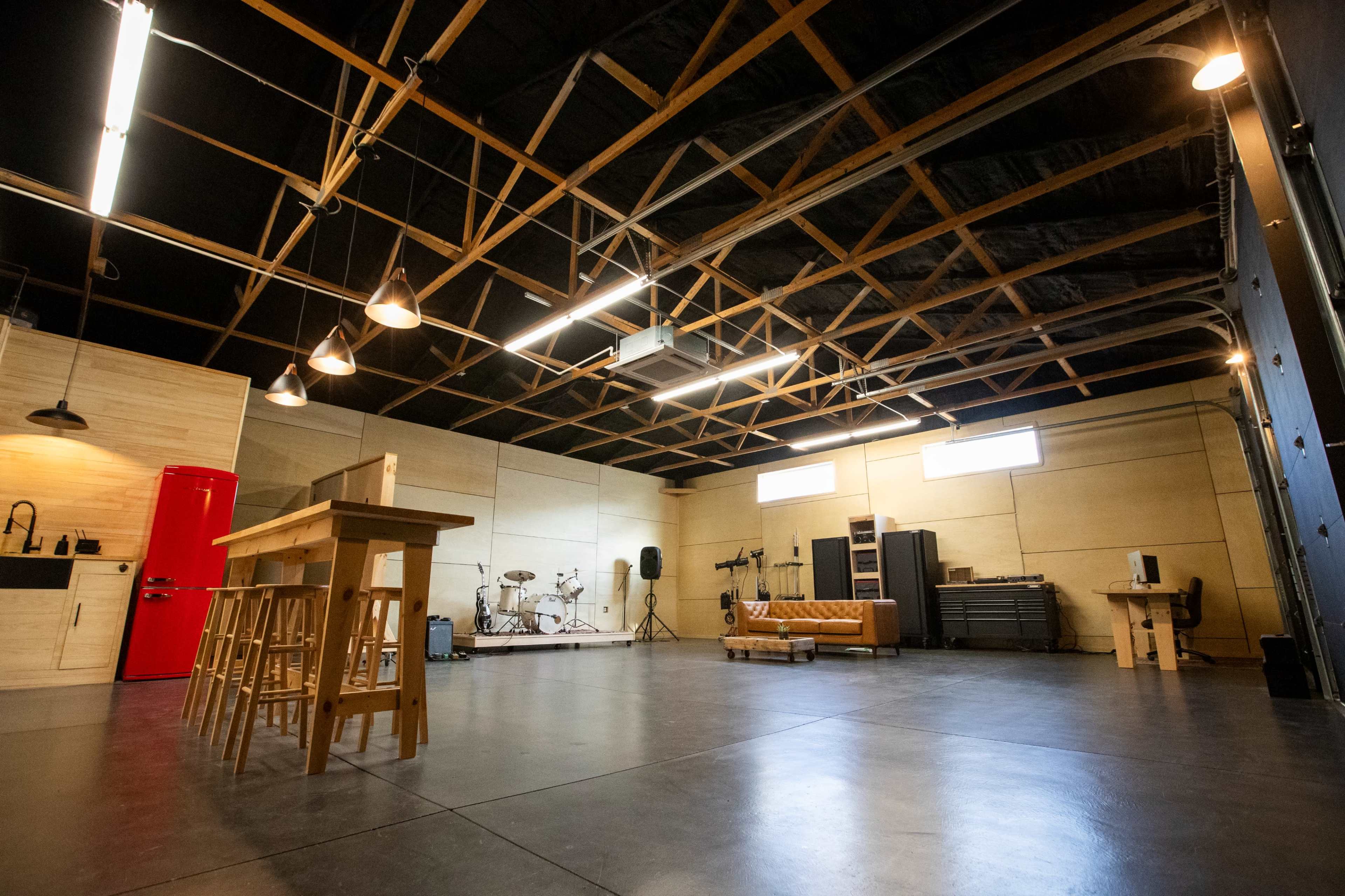 The image shows a spacious studio with a hardwood ceiling, equipped with a kitchen area, musical instruments, sound equipment, and a large couch.