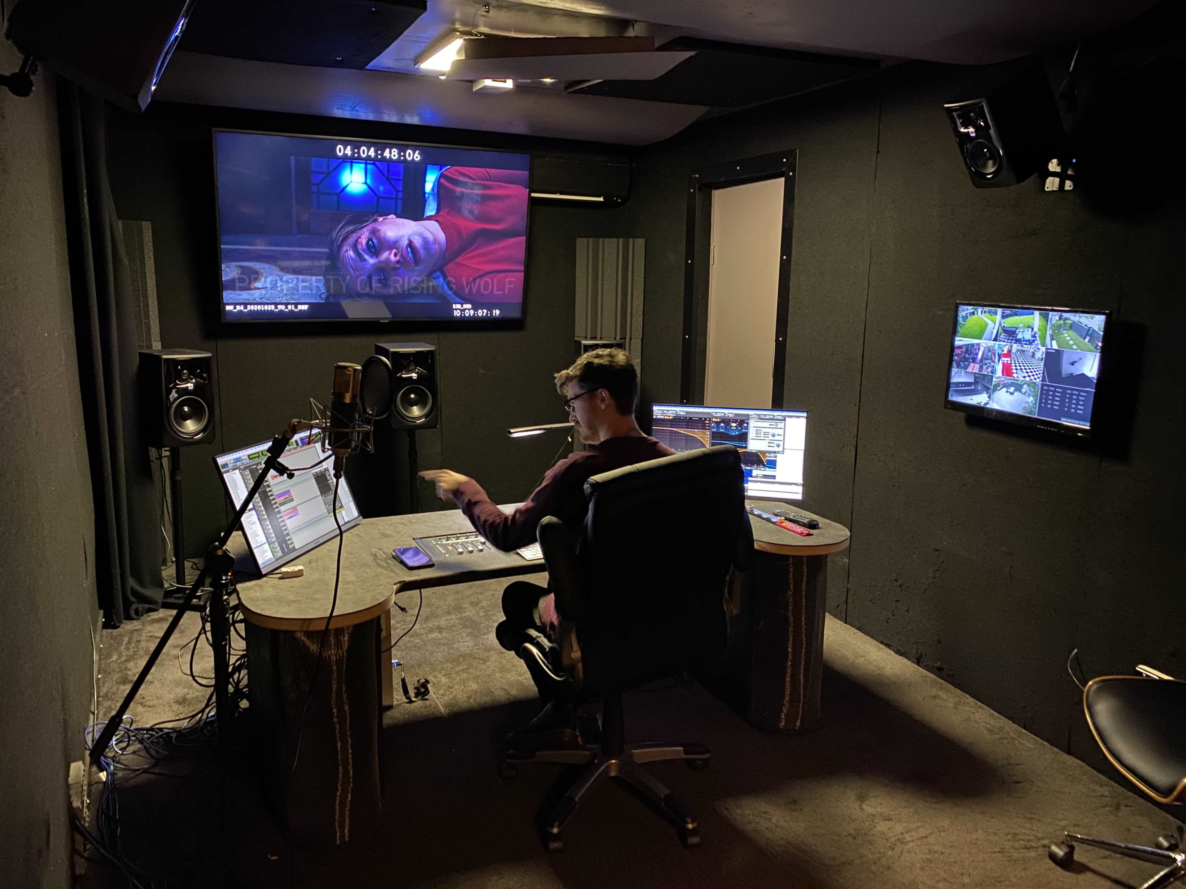 A person is seated at a computer desk in a recording studio, surrounded by monitors and audio equipment while working on a project.