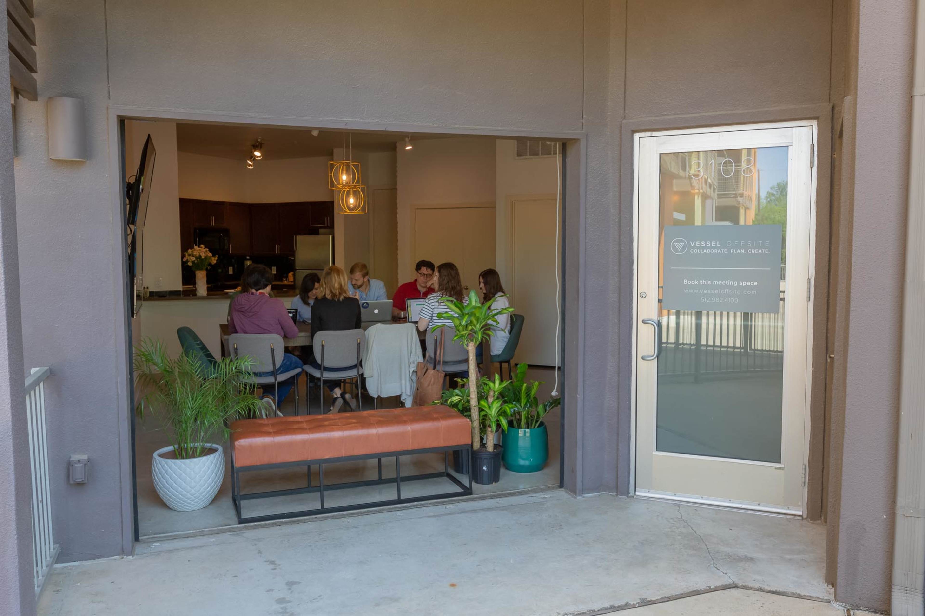 A group of six people is seated around a table in a modern office meeting space, visible through a large glass door.