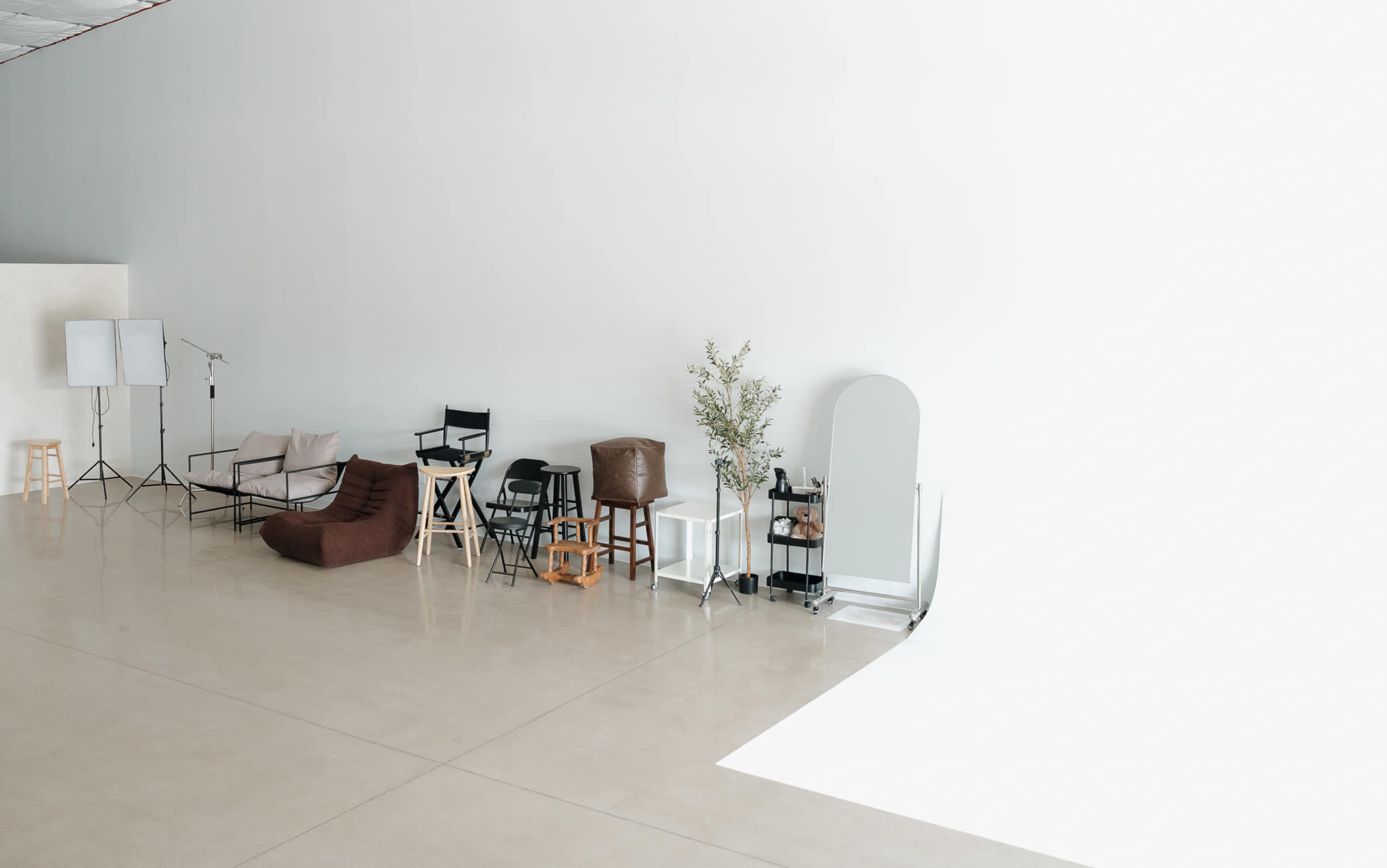 The image shows a spacious studio interior featuring various chairs, a mirror, and a plant arranged along a light-colored wall.