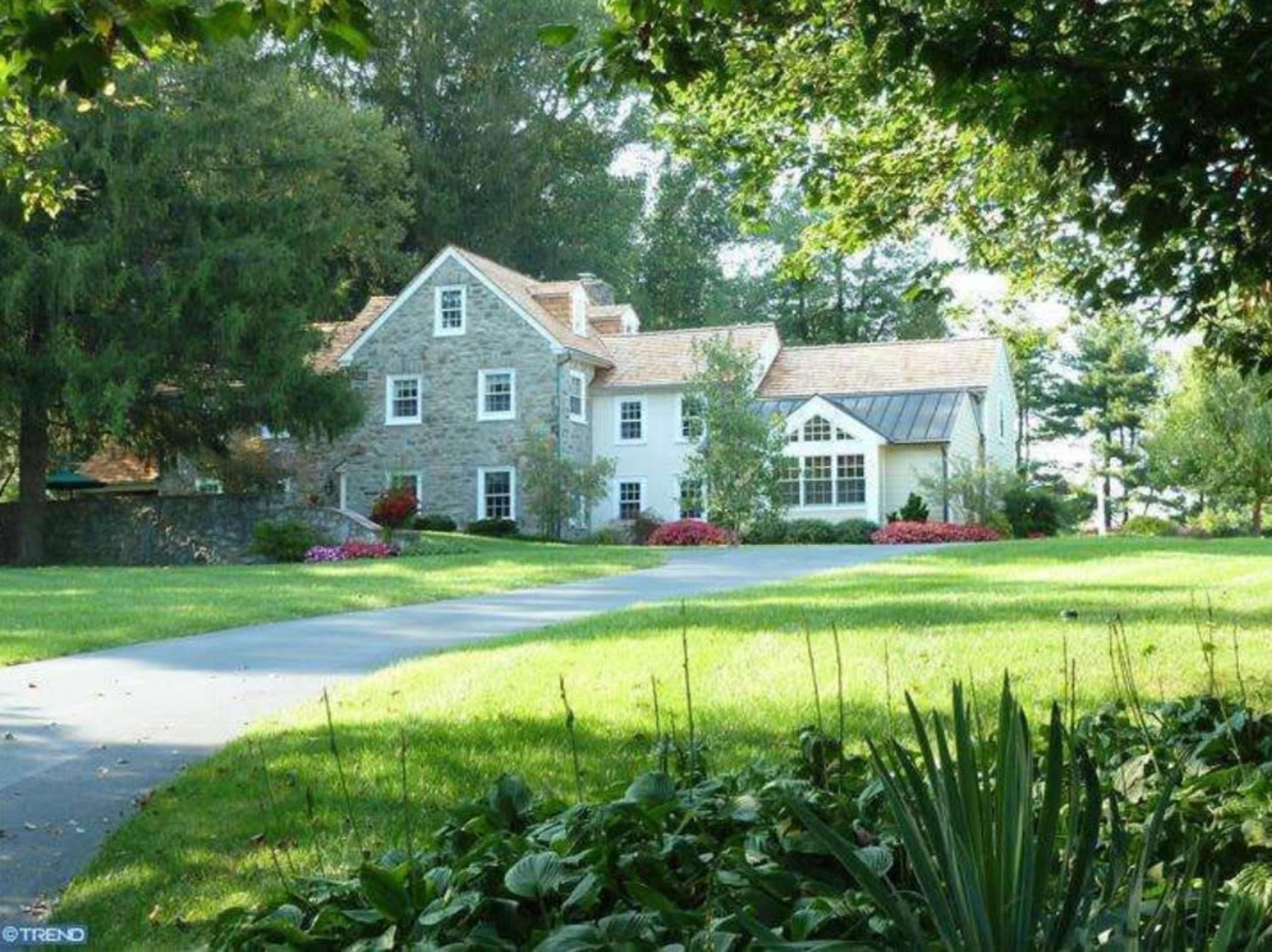 A stone house with a landscaped yard and a long driveway is surrounded by trees and flowers.
