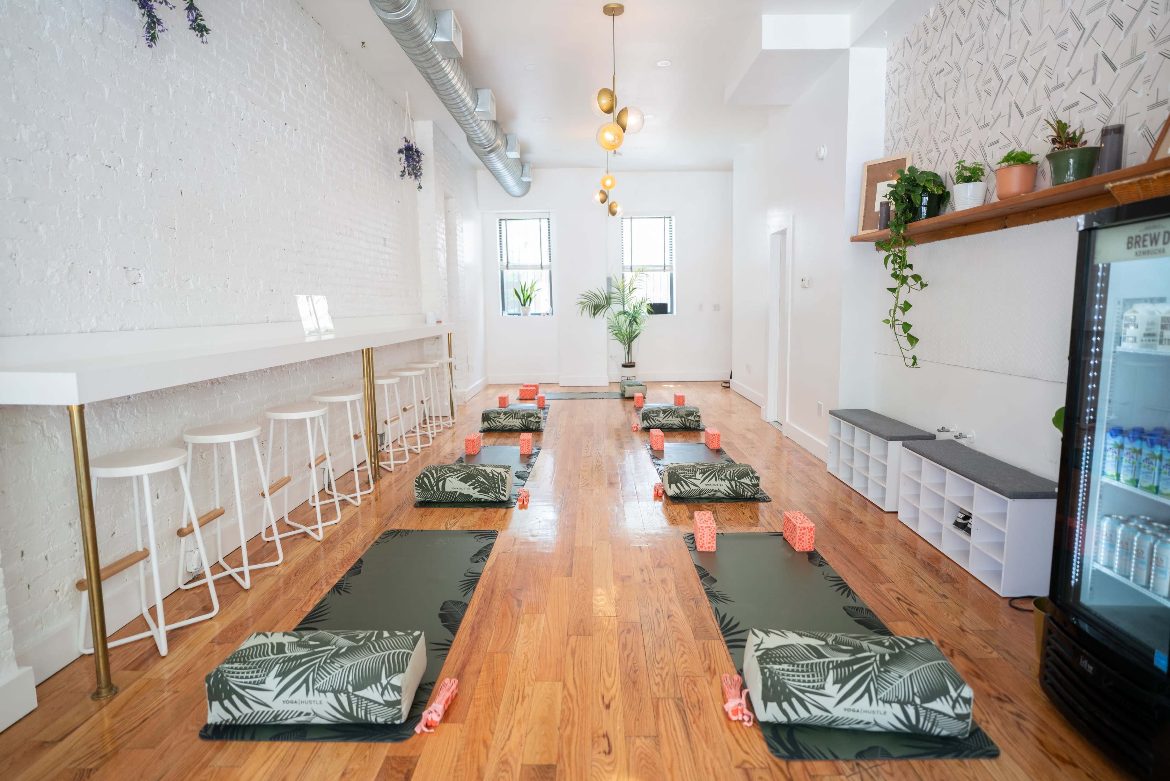 A yoga studio with several mats arranged on a wooden floor, a seating area with bar stools, and plants decorating the space.