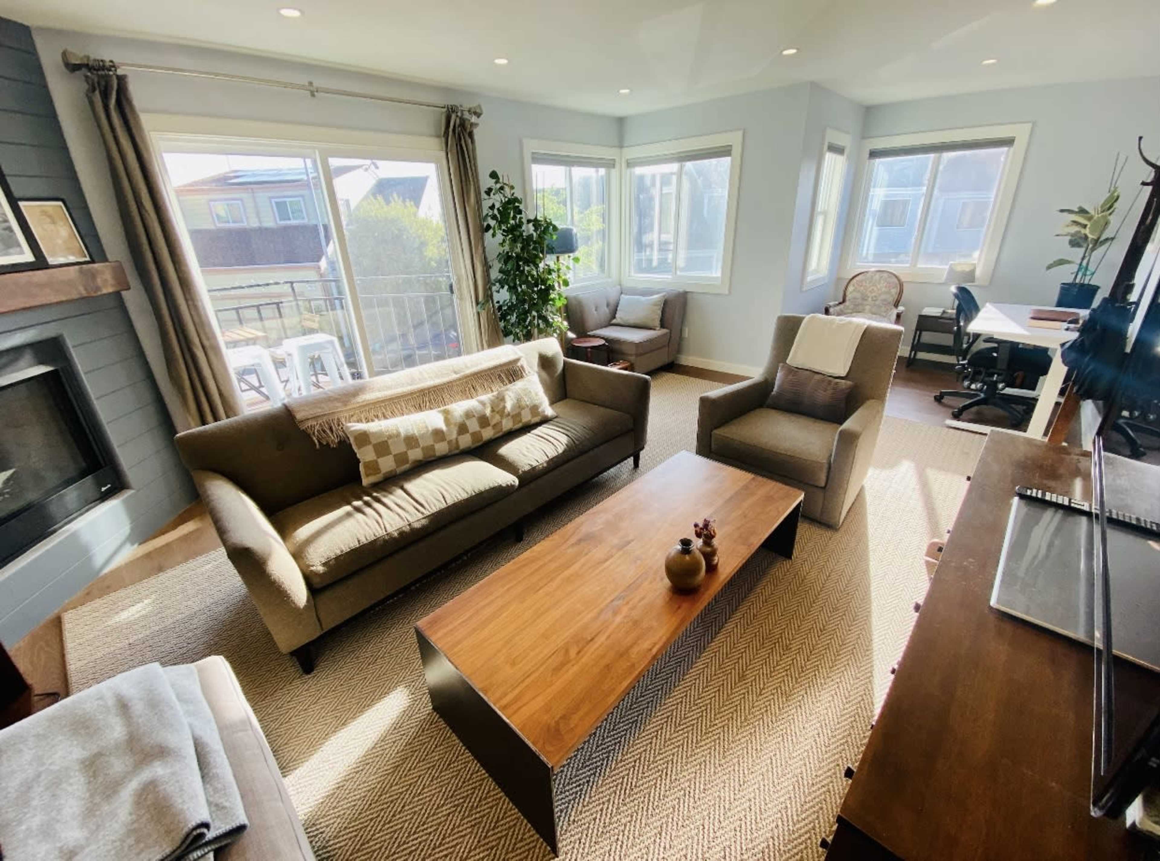 A well-lit living room with a sofa, armchairs, a coffee table, and large windows overlooking an outdoor area.
