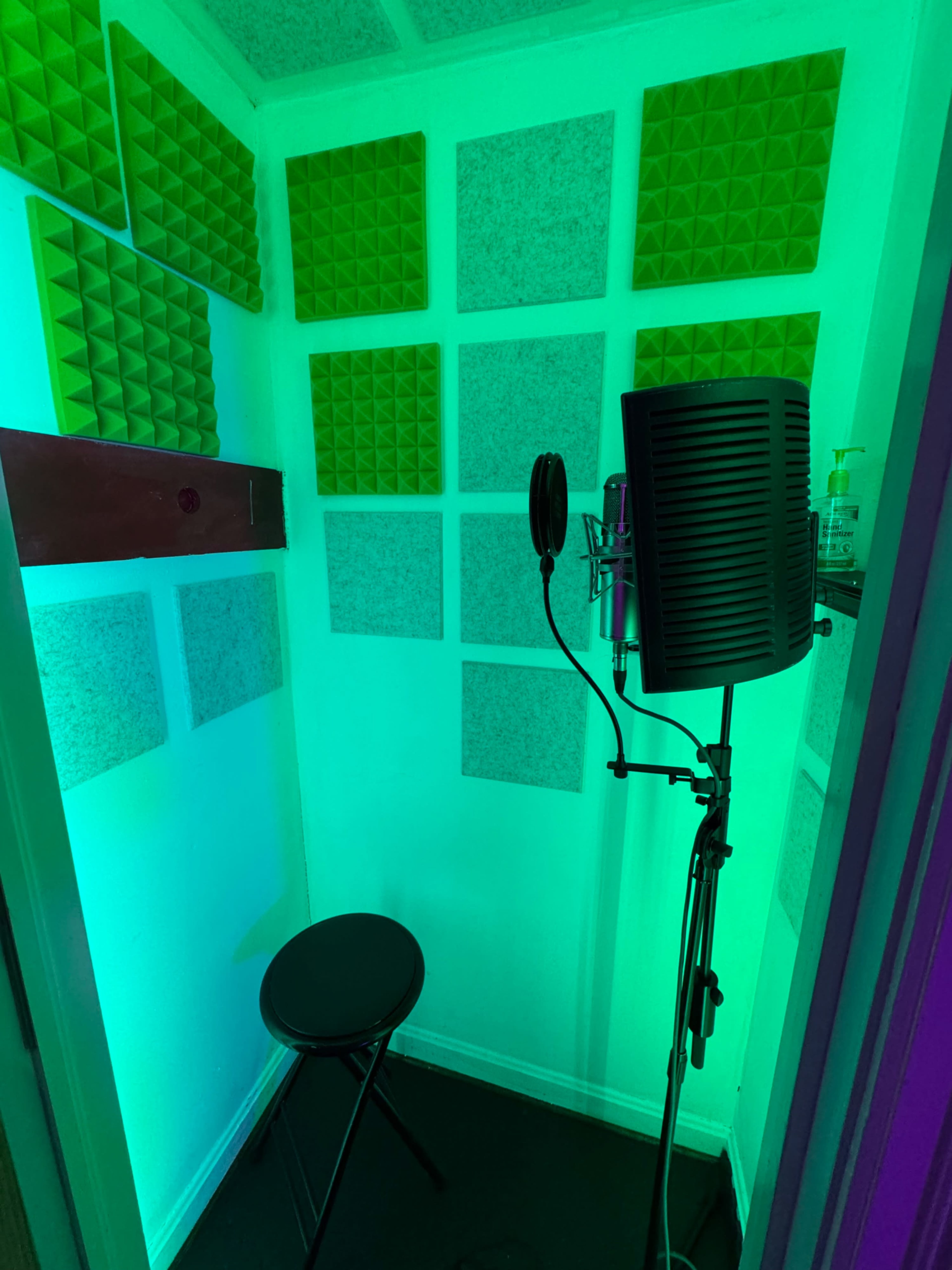 The image shows a small soundproof recording booth with green acoustic foam panels on the walls, a microphone on a stand, and a black stool in the corner.