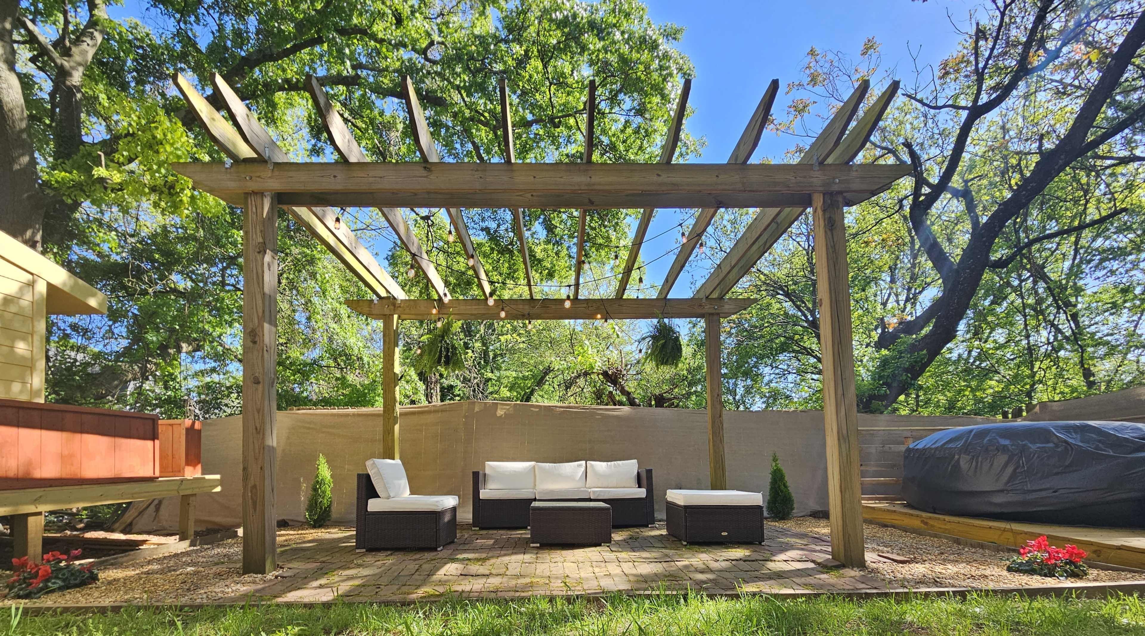 A wooden pergola with a set of outdoor seating surrounds sits in a landscaped yard under a clear blue sky.