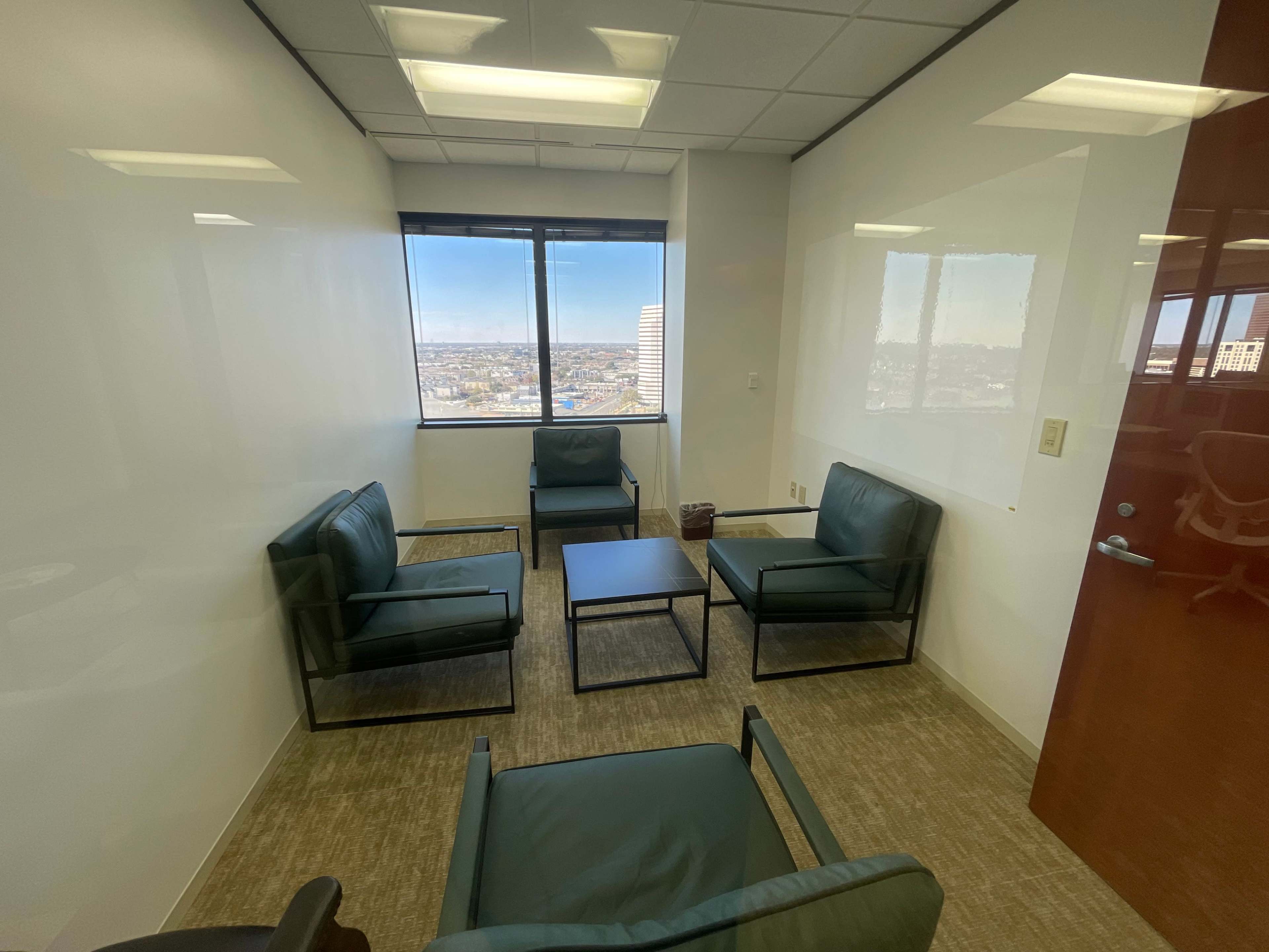 The image shows a small meeting room with four green chairs arranged around a black table, featuring large windows that overlook a cityscape.