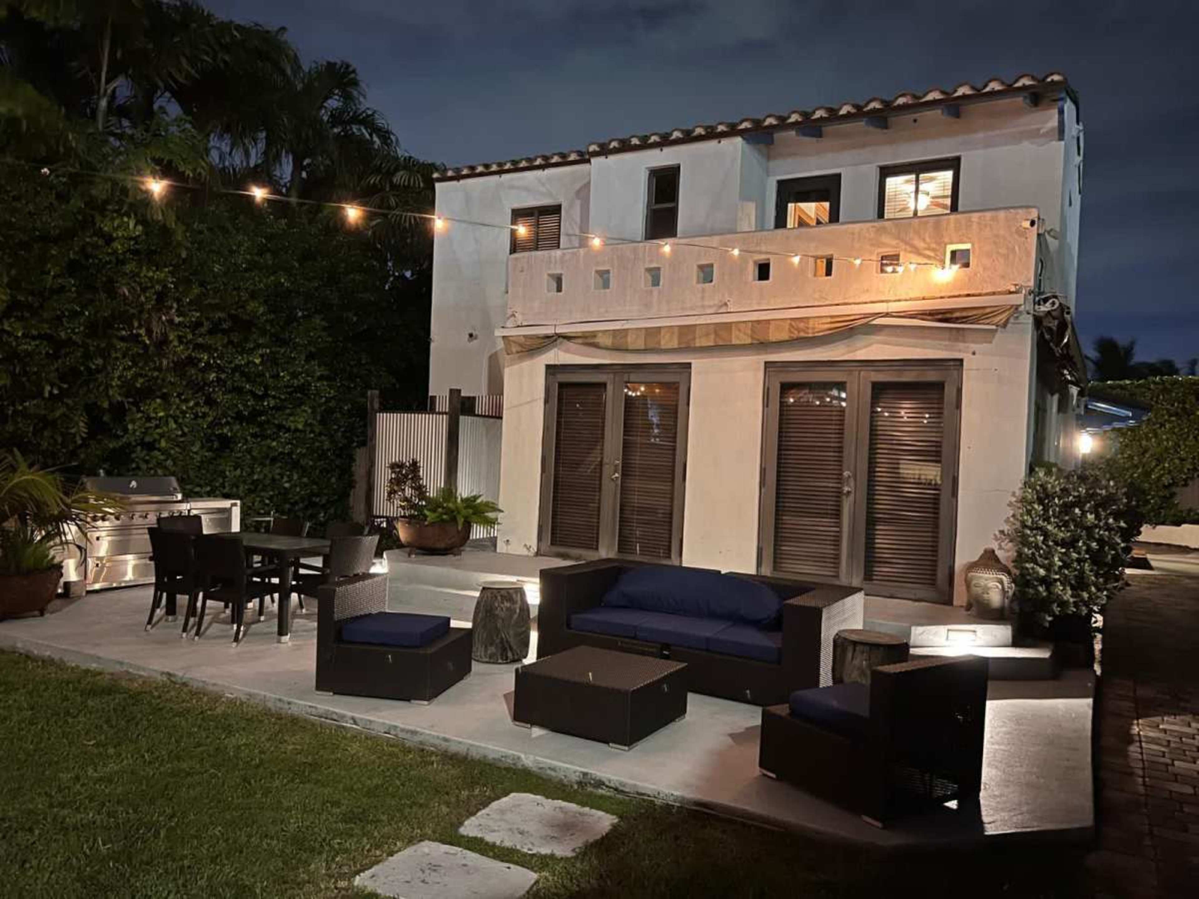 A modern two-story house with outdoor seating and a dining area illuminated by string lights in the evening.